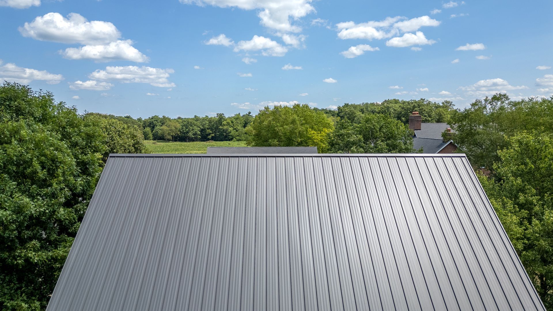 Metal roof under a blue sky with clouds, surrounded by green trees and a glimpse of a house.