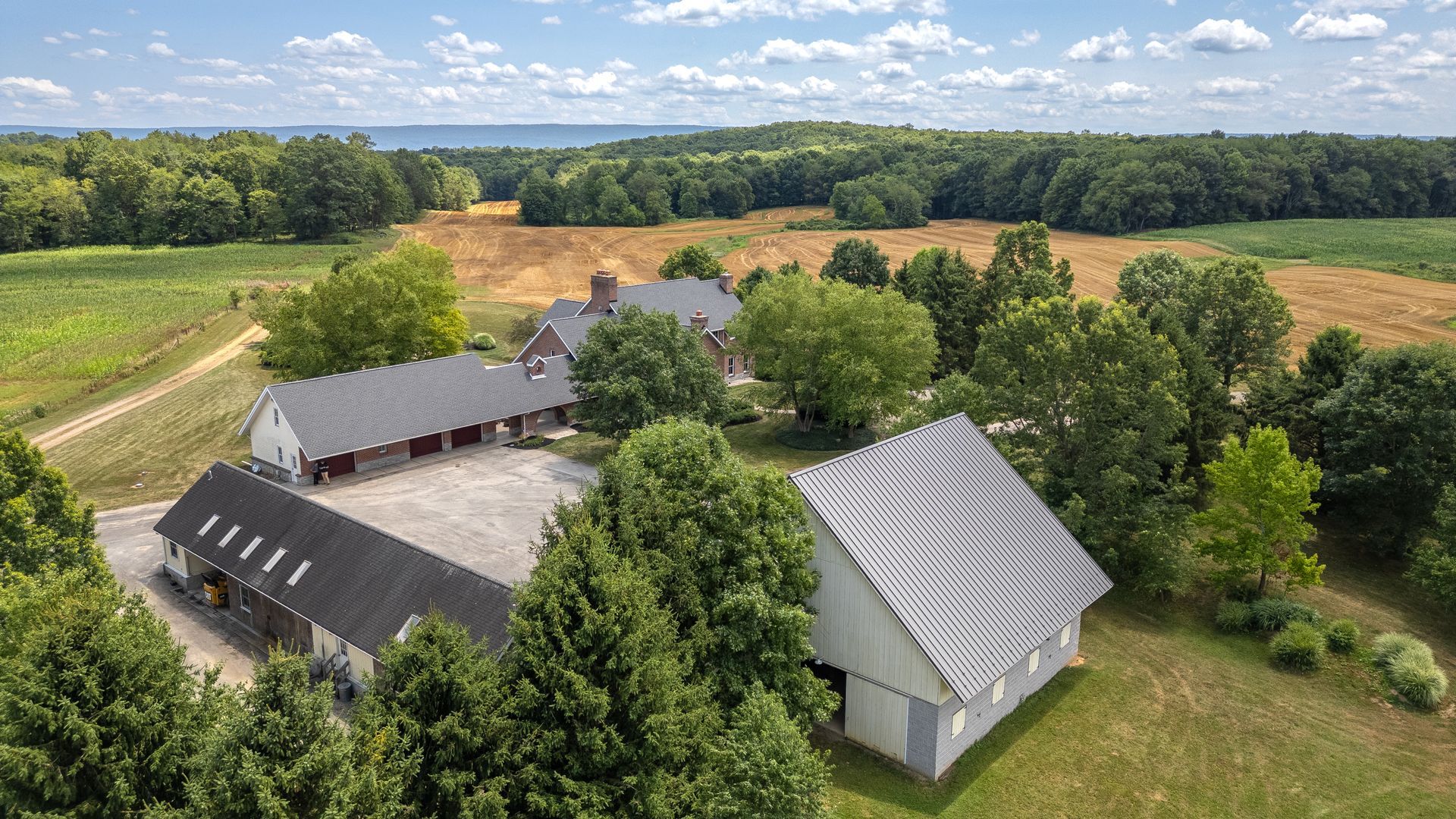 Aerial view of a farm with multiple buildings, fields, and trees. Sky with clouds in the background.