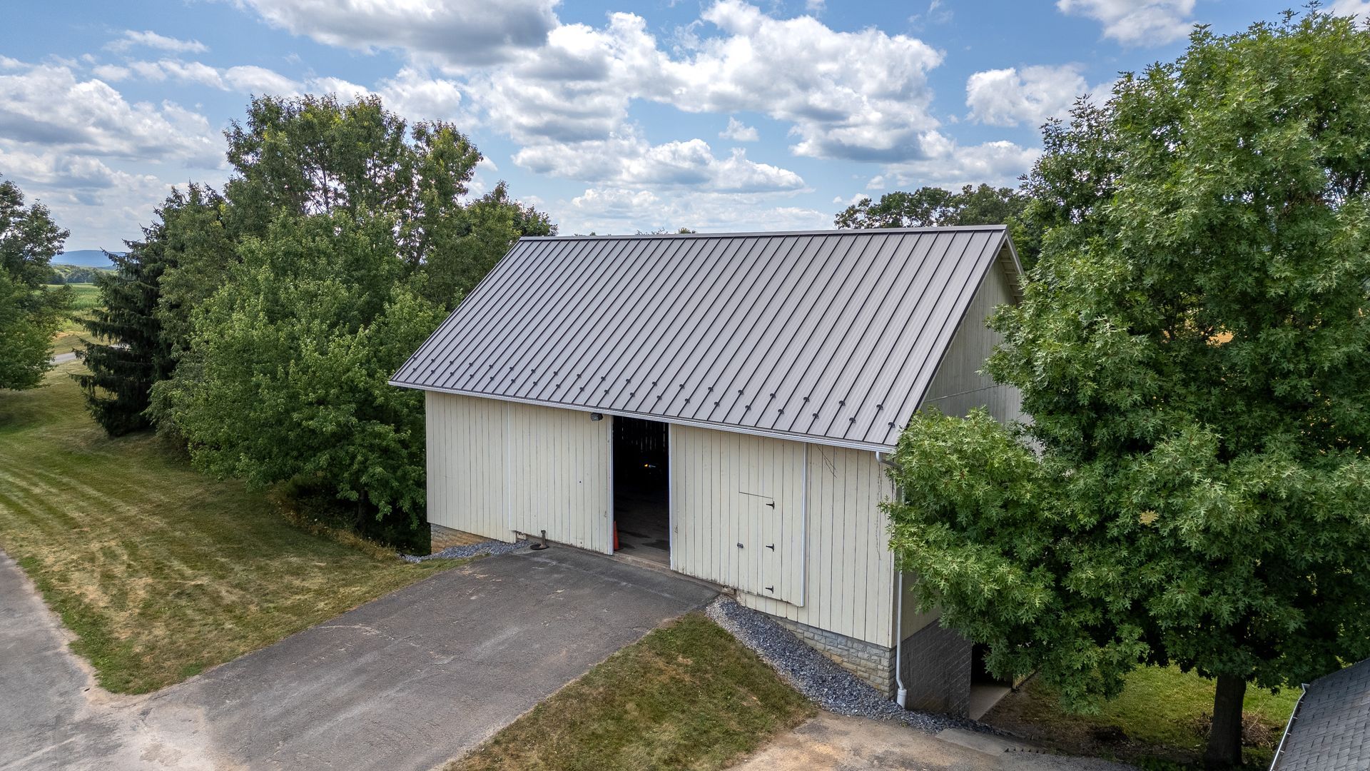 White barn with gray roof, trees, and driveway under a cloudy sky.