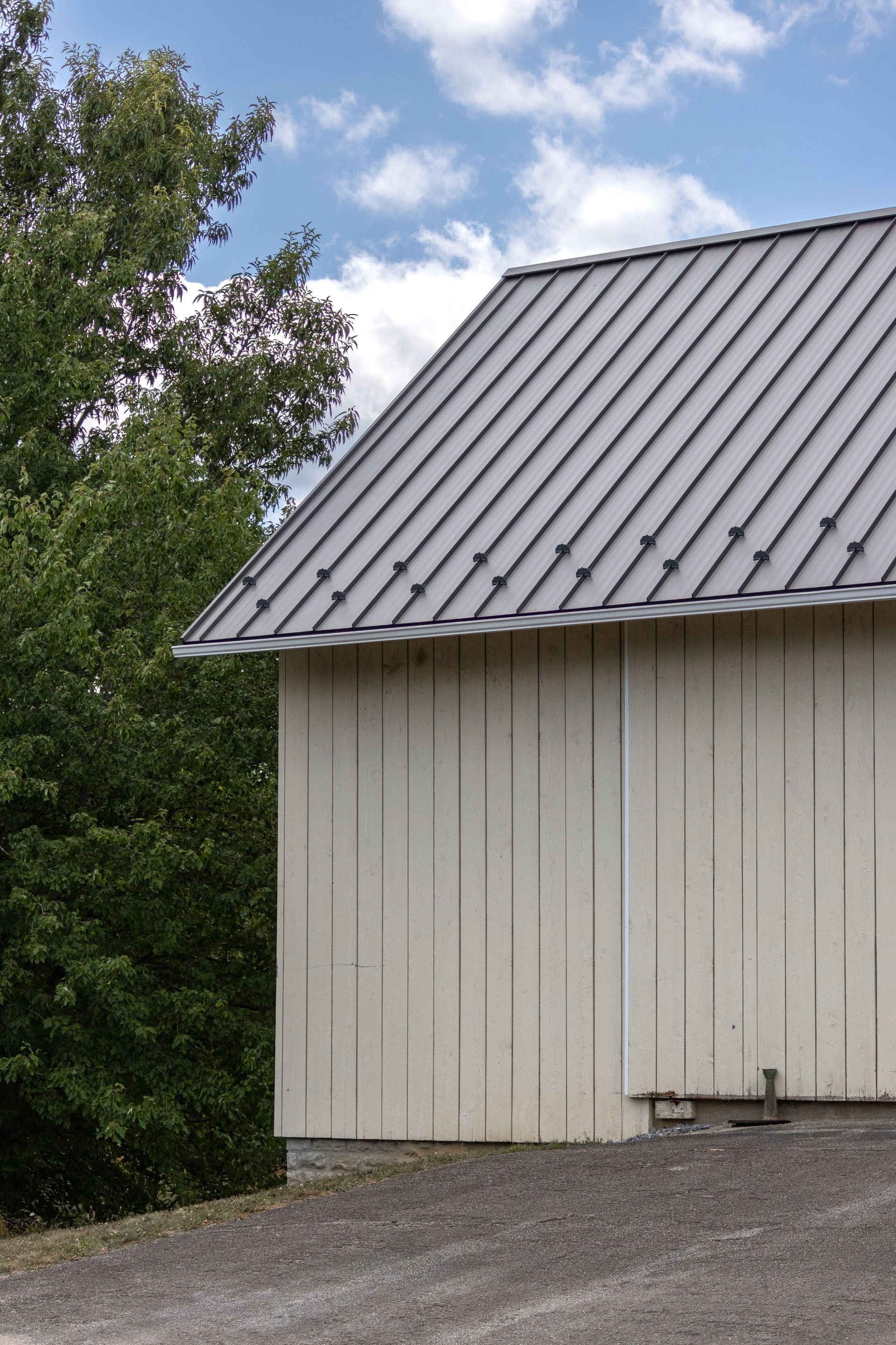 White barn with gray metal roof, next to a tree, gravel ground, blue sky with clouds.
