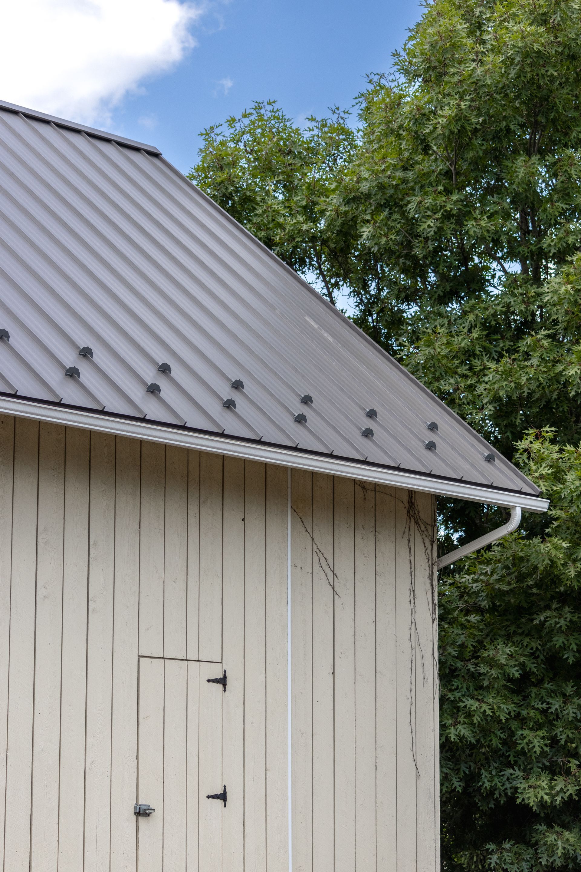 Beige barn with a gray metal roof and a white gutter, trees in the background.
