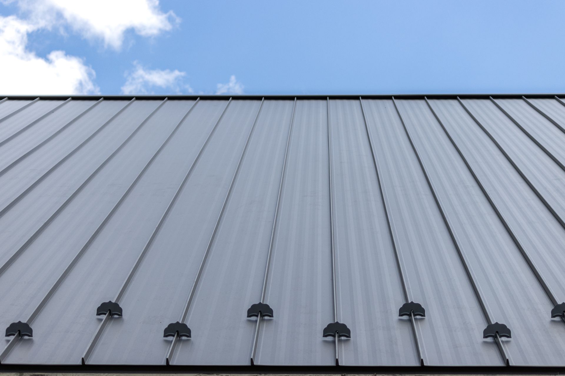 Silver metal roof panels with black fasteners against a blue sky with scattered clouds.