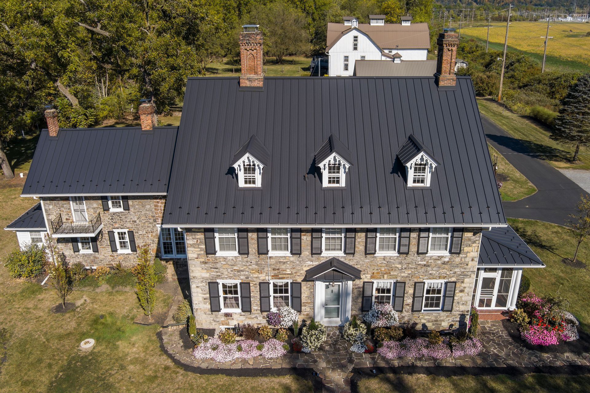 An aerial view of a stone house with a black metal roof, three gabled dormers, and dark shutters, surrounded by gardens.