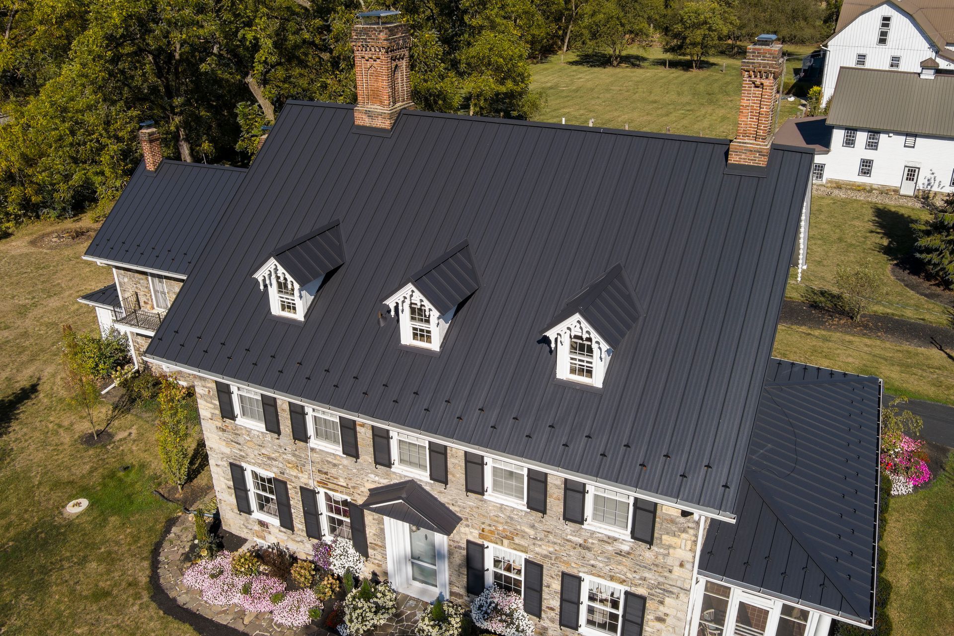 An aerial view of a stone house with a dark, textured metal roof, three dormer windows, and two brick chimneys.