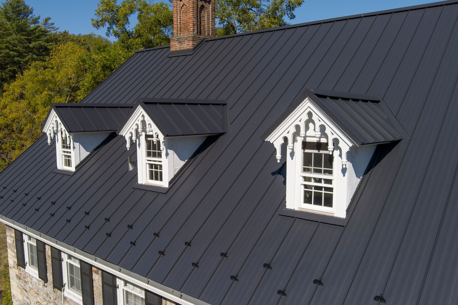 A dark standing-seam metal roof with three white dormers featuring ornate trim on a stone building.