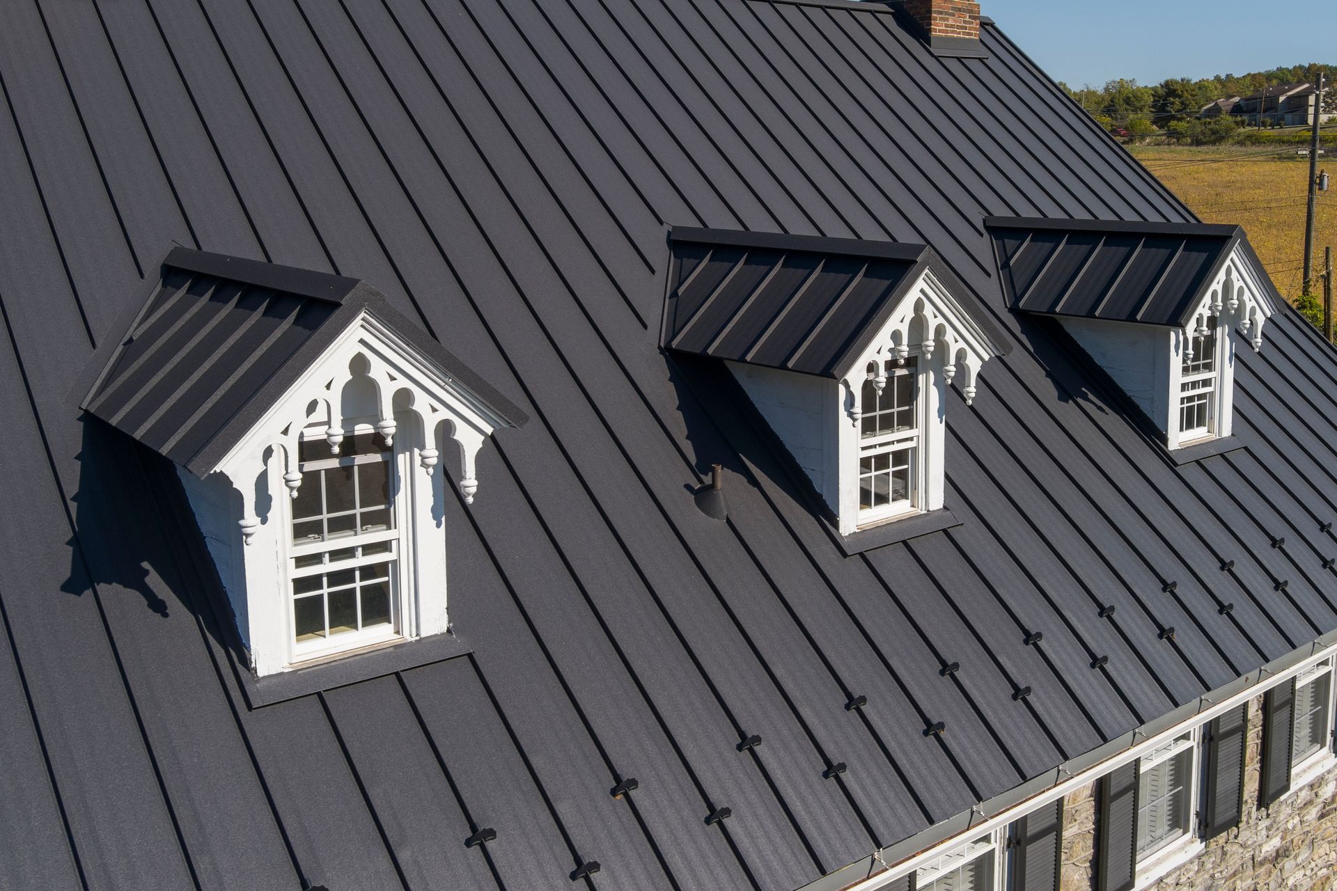 A dark metal roof features three white, ornate dormer windows with decorative woodwork against a bright blue sky.