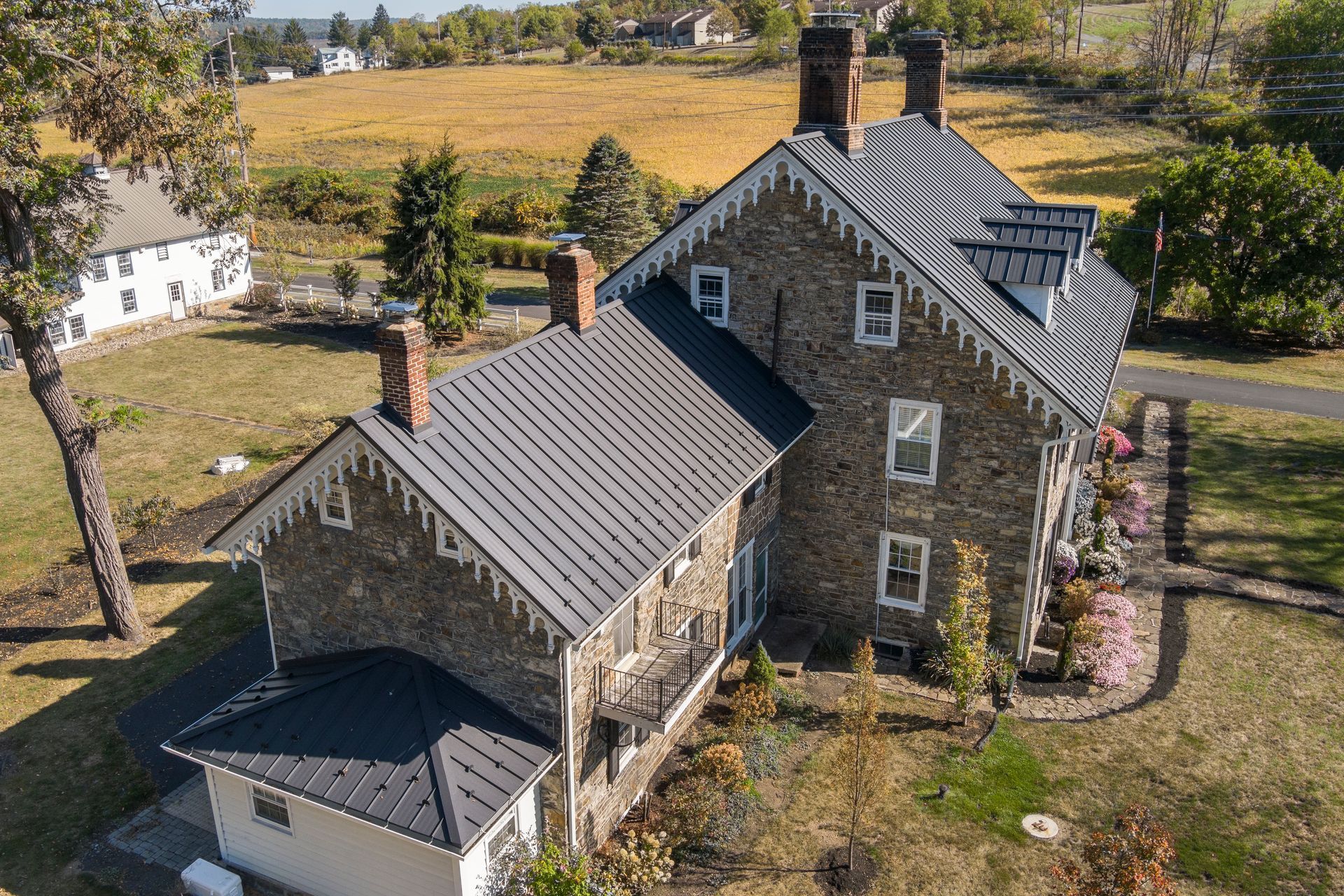 An aerial view of a historic two-story stone house with a dark metal roof and ornate white trim, surrounded by a rural field.