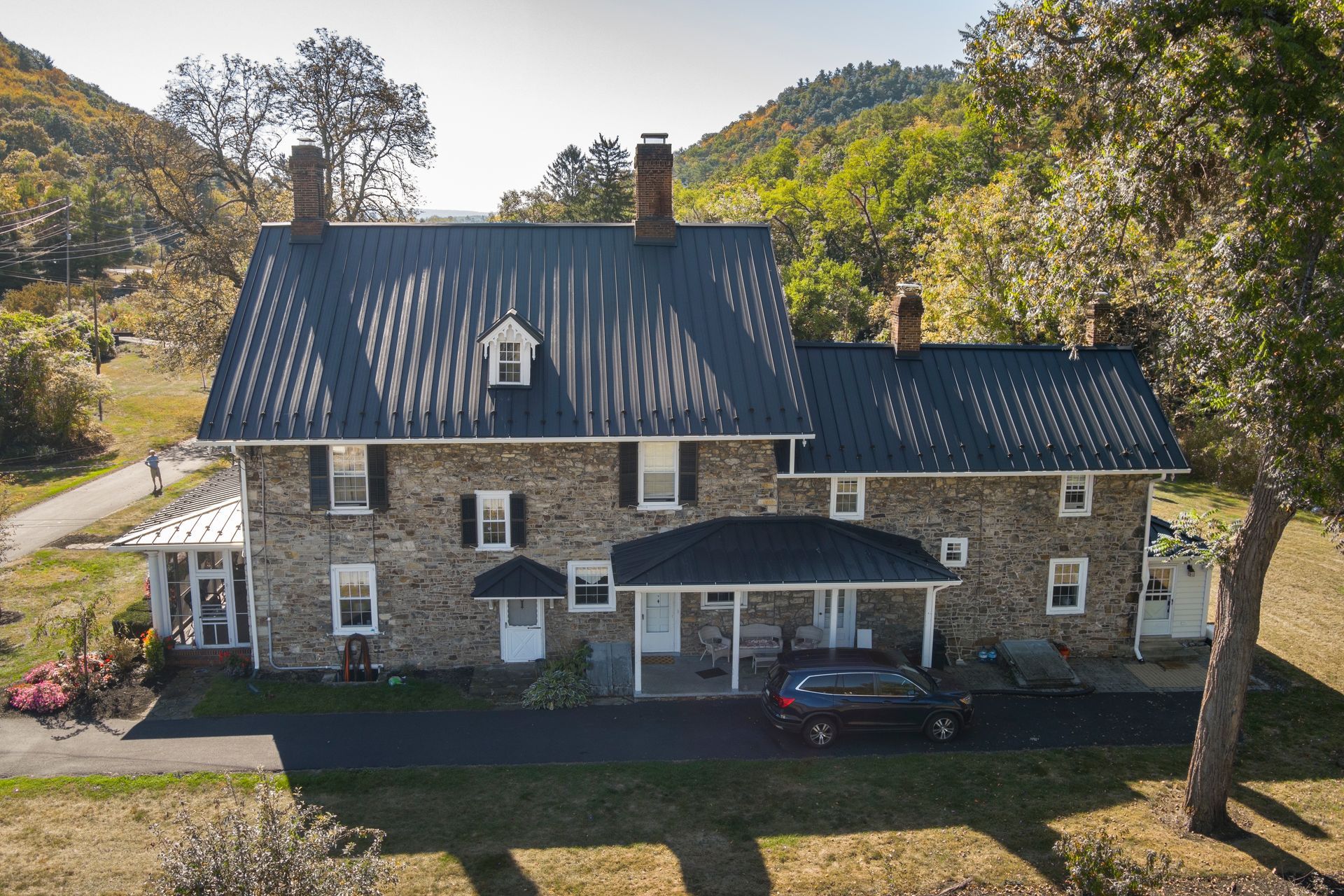 A stone farmhouse with a dark metal roof, surrounded by a rural landscape with wooded hills in the background.