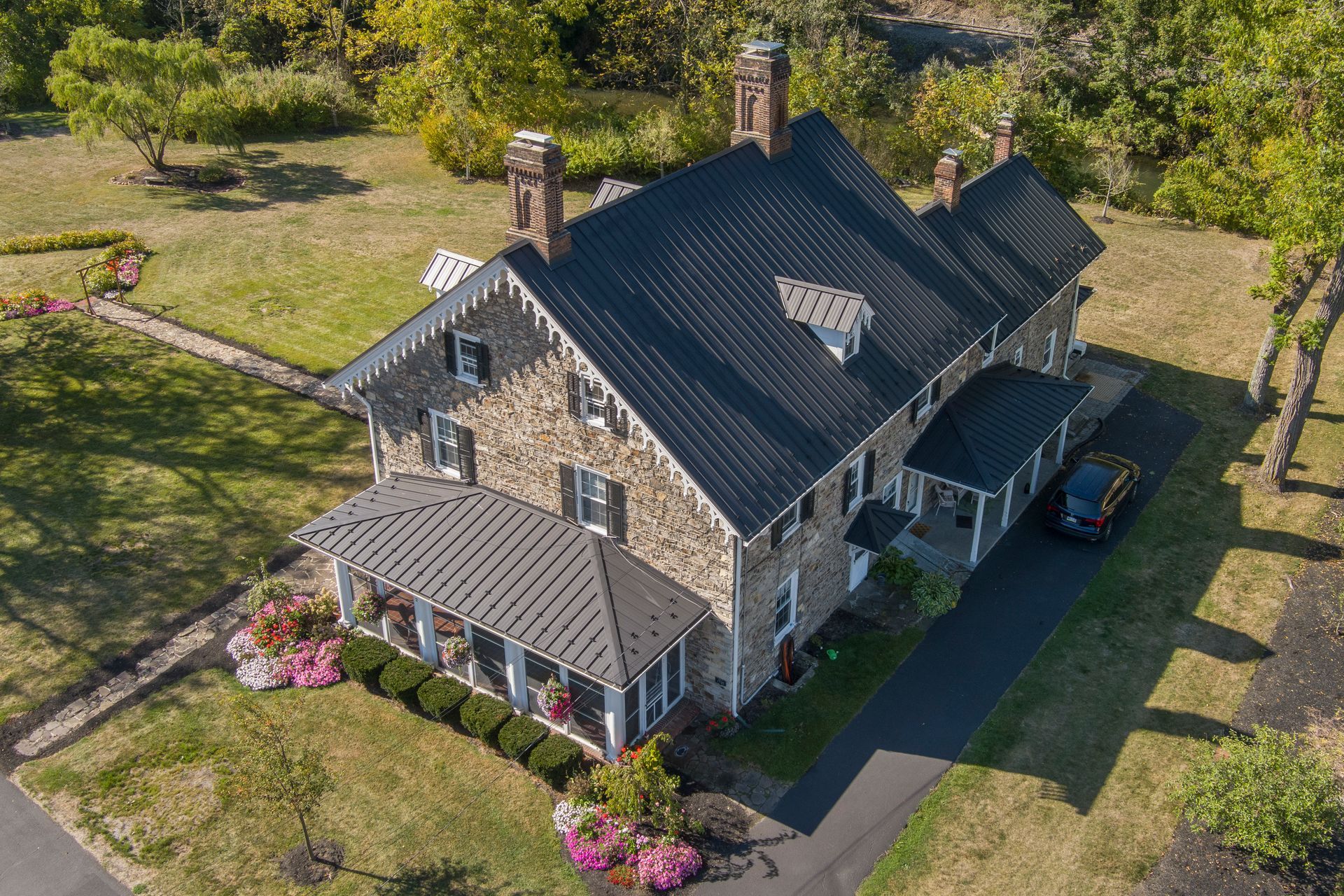 An aerial view of a stone house with a dark metal roof, surrounded by a lawn, garden beds, and a paved driveway.