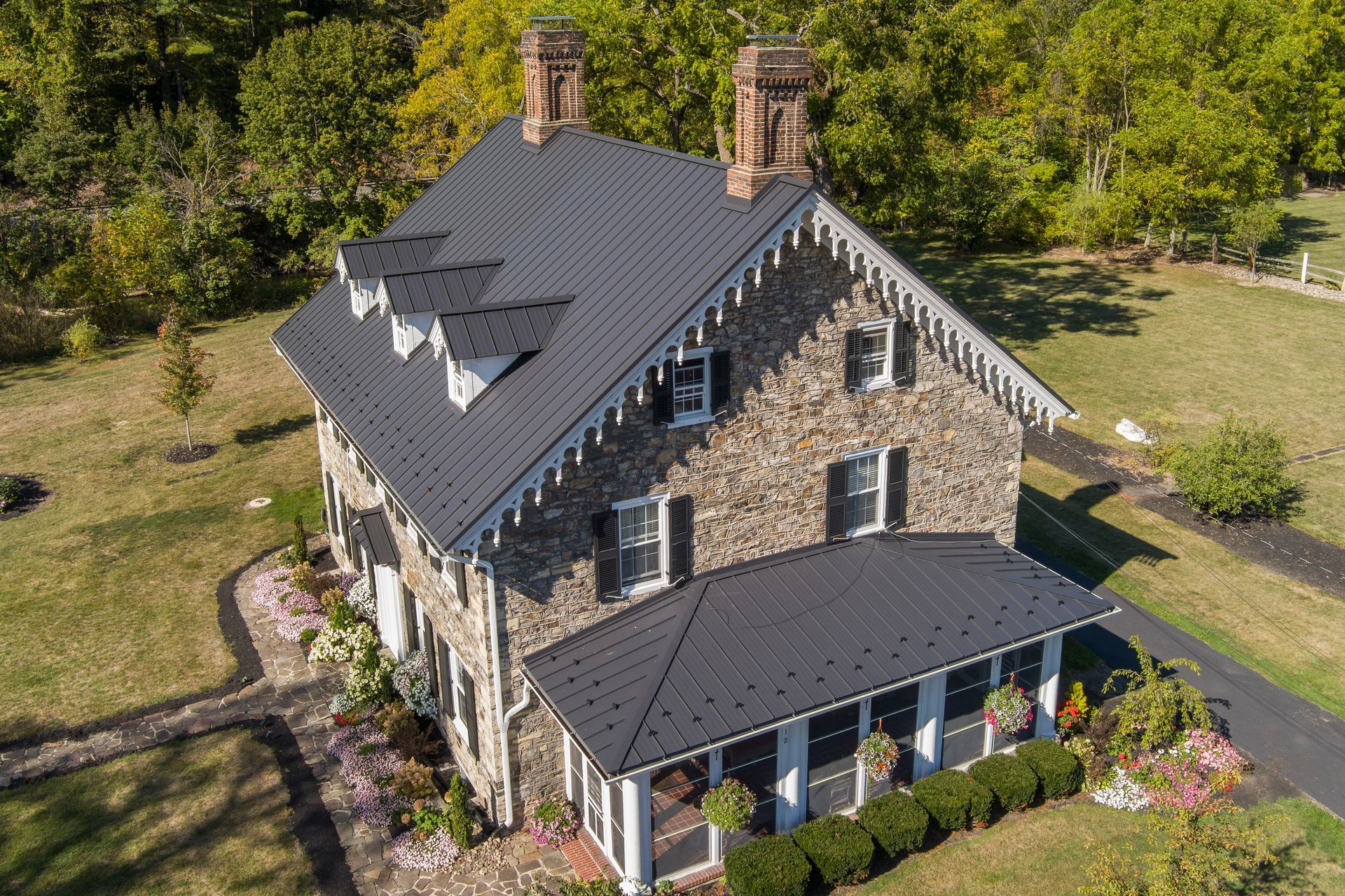 An aerial view of a stone house with a dark metal roof, set on a grassy lawn with trees in the background.