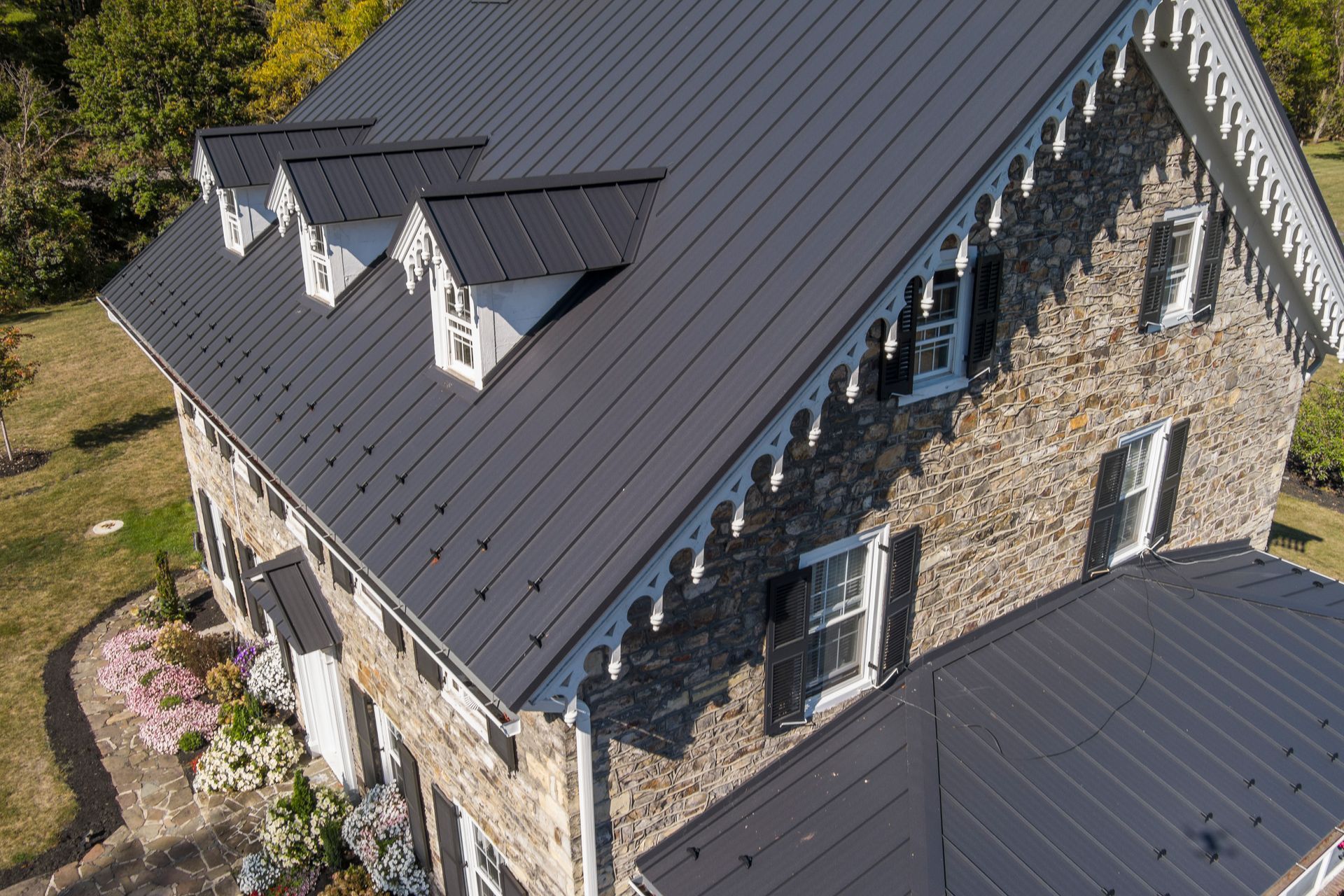A stone house with a dark metal roof, decorative white trim, and multiple dormer windows, viewed from above.