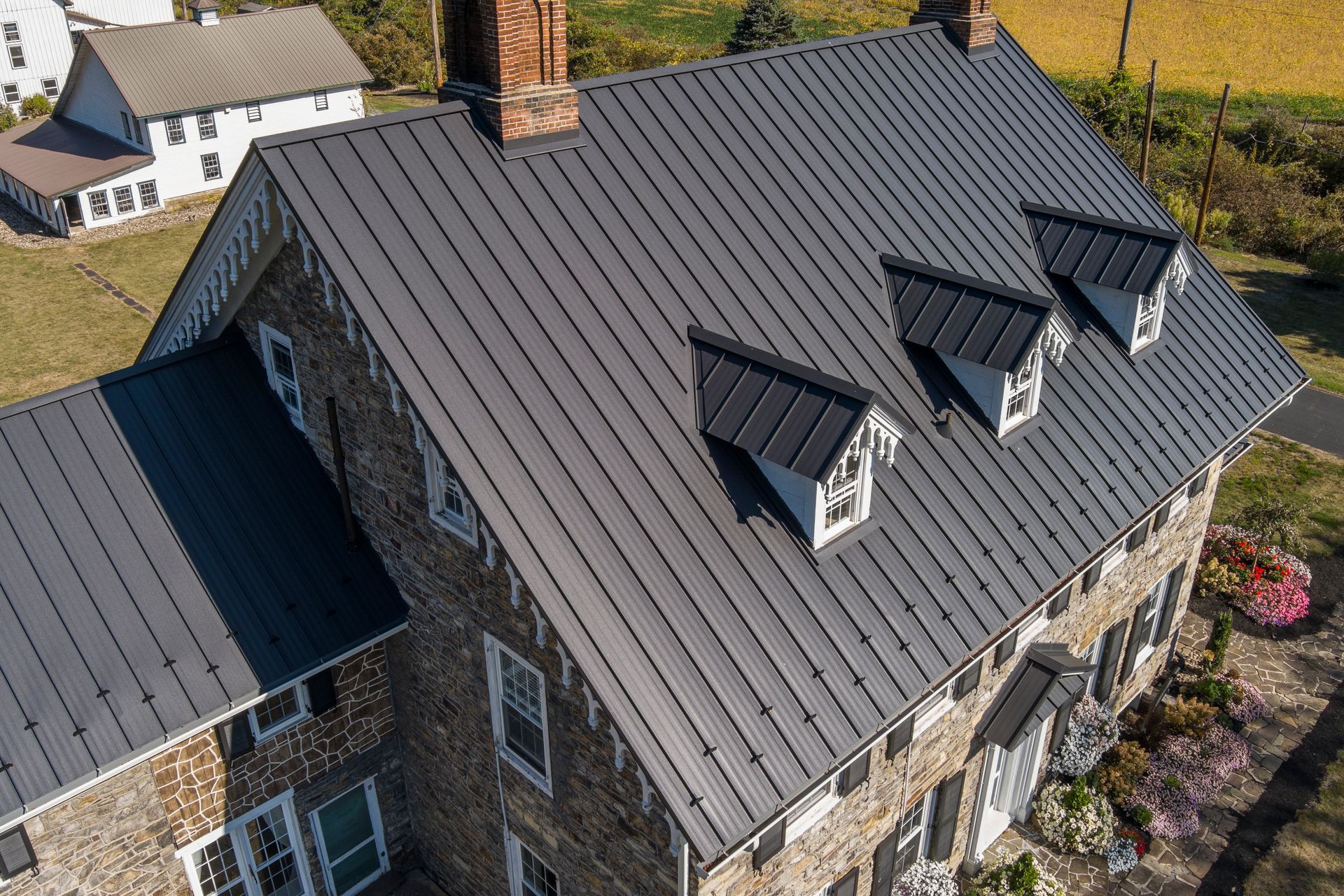 A stone house with a dark metal roof, three small dormers, and a brick chimney, seen from an aerial angle.