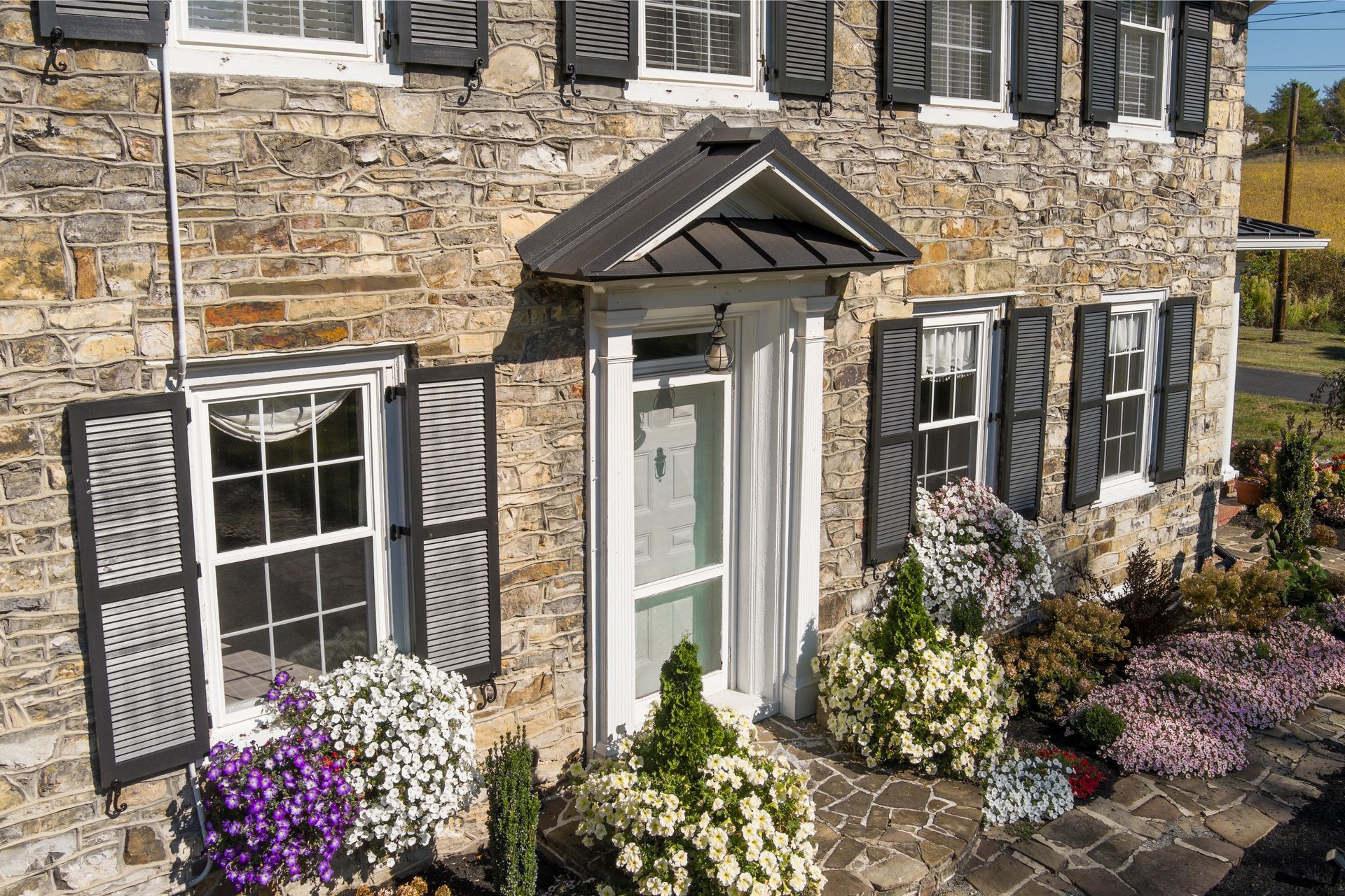 A stone cottage front with a black peaked porch roof, dark shutters, and flower-filled garden beds in front.