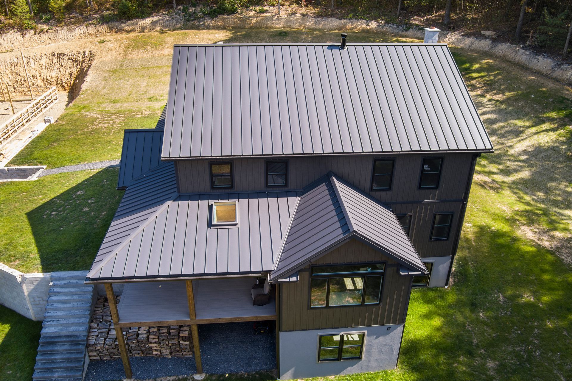 An aerial view of a dark-sided house with a black metal roof, featuring a deck, stone landscaping, and a grassy yard.
