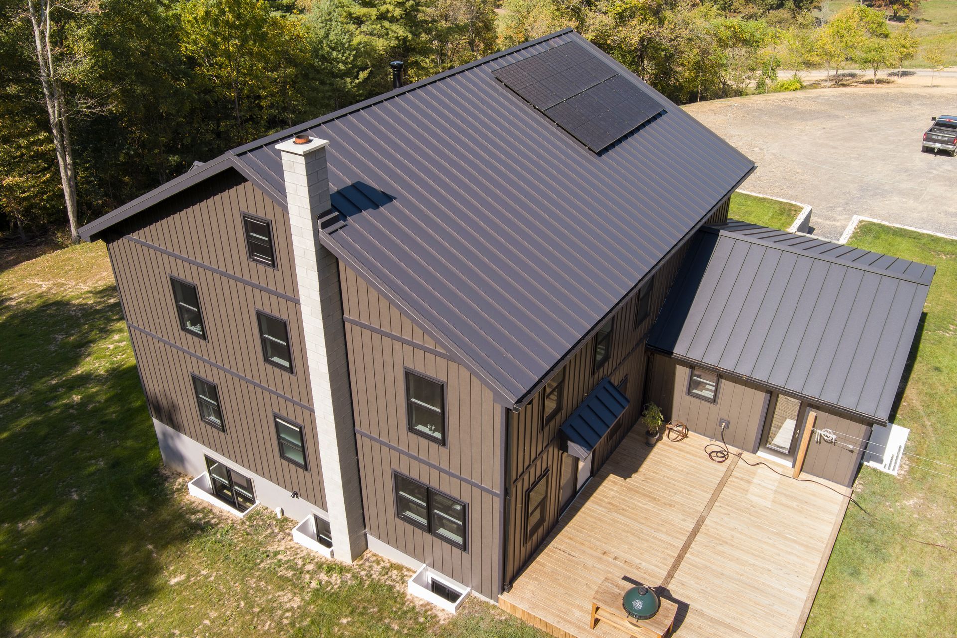 An aerial view of a dark brown, two-story modern house with a black metal roof, solar panels, and a wood patio.