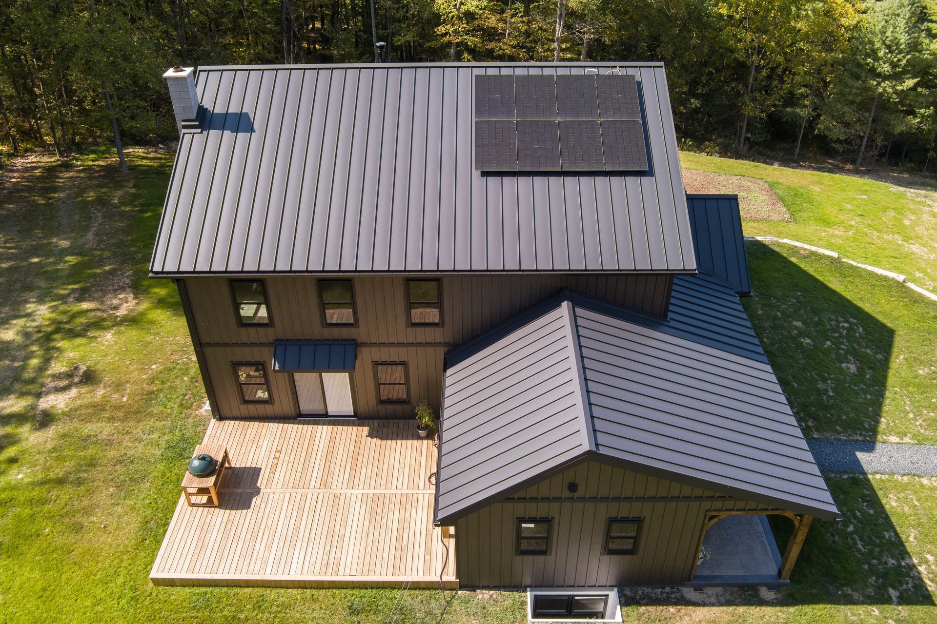 Aerial view of a brown two-story house with a black metal roof, solar panels, and a wood patio in a wooded yard.