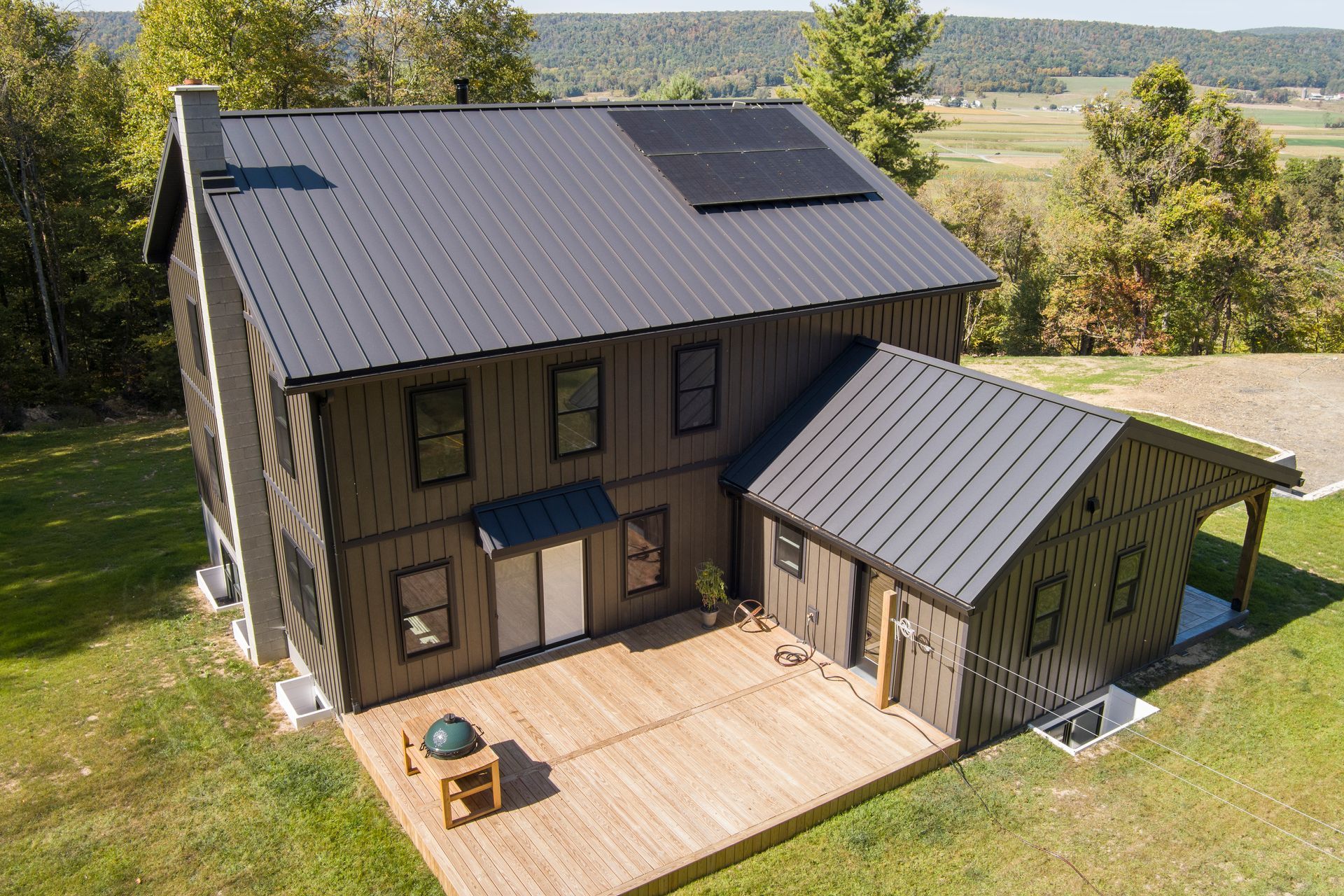 An aerial view of a two-story home with dark metal siding and a matching gabled roof, featuring a deck and solar panels.