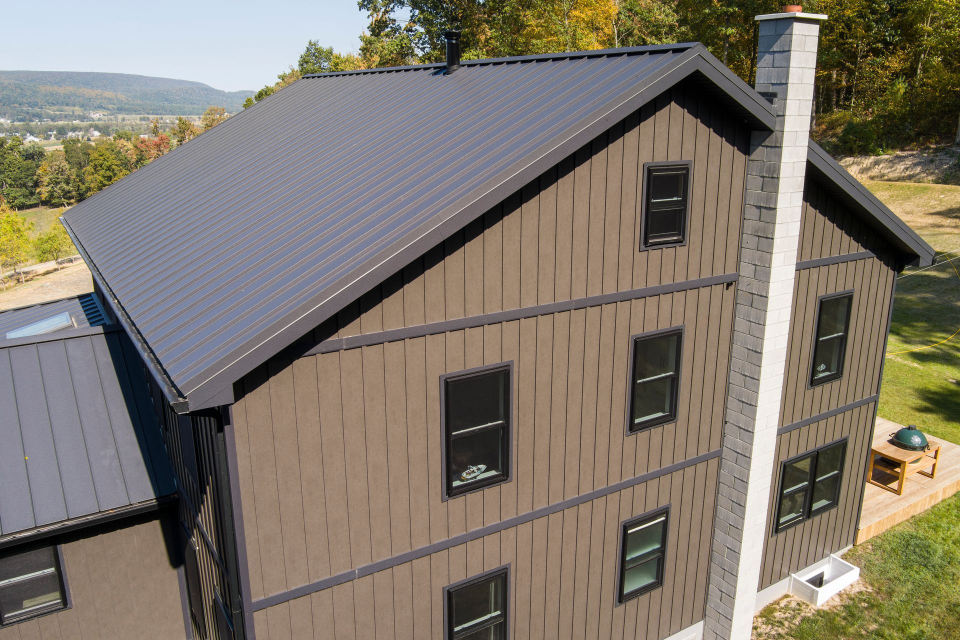 A three-story house with dark metal roofing, brown textured panel siding, black-framed windows, and a tall stone chimney.