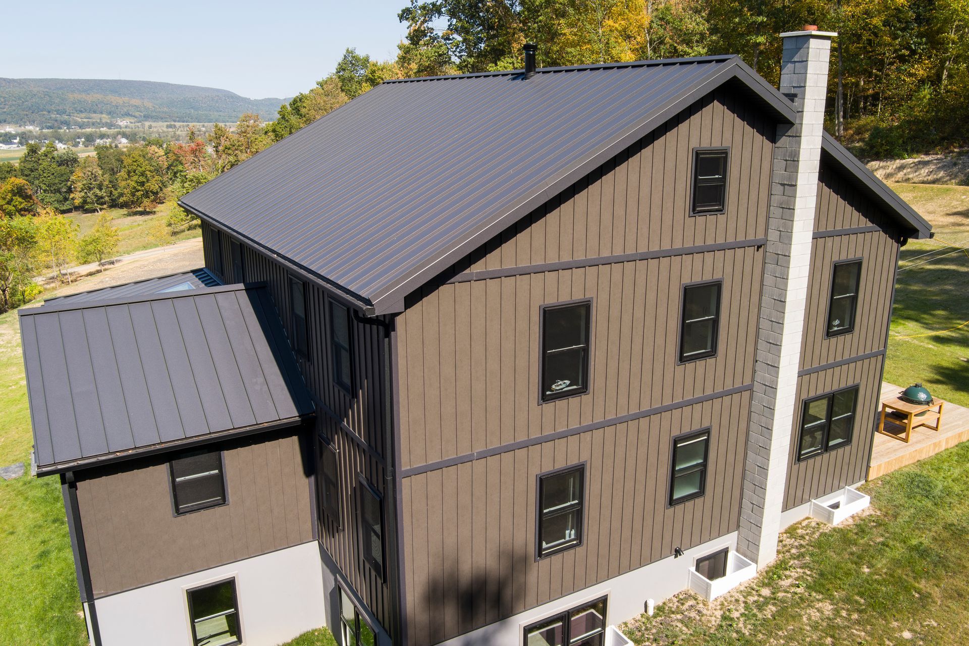 An aerial view of a multi-story house under construction with dark brown panels, black metal roof, and a stone chimney.