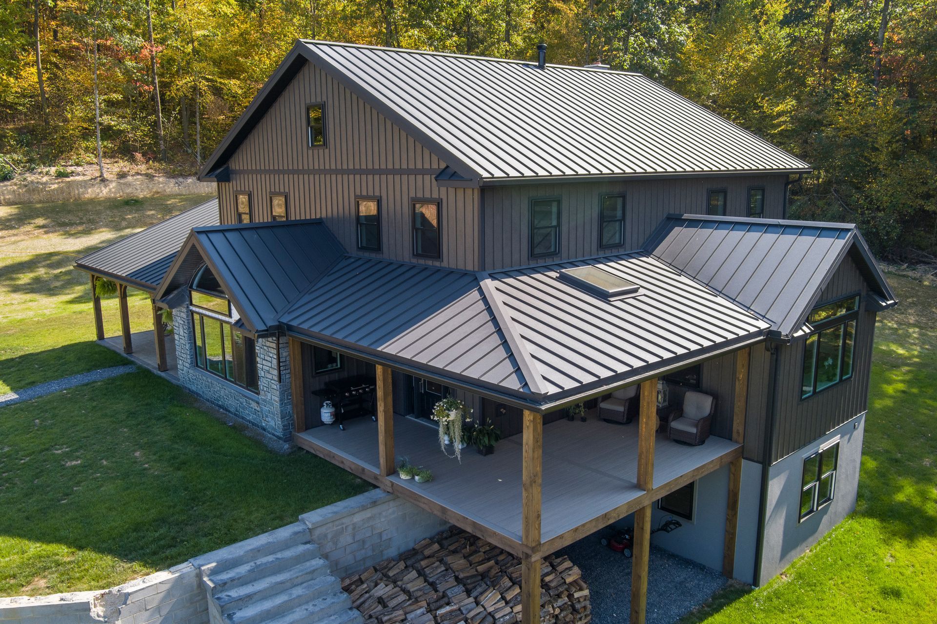 A two-story dark-sided house with a black metal roof, a wrap-around porch, and a stone foundation set in a wooded area.