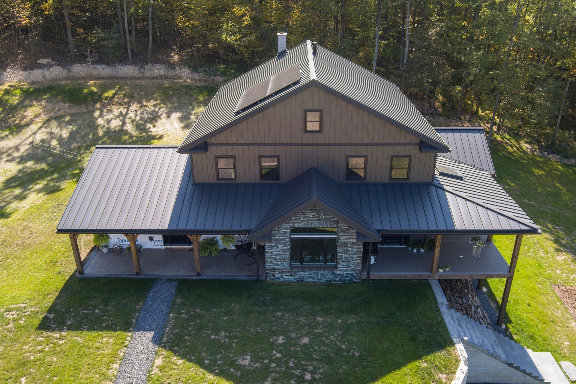 A two-story brown house with a dark metal roof, stone entry, and wrap-around porch, nestled in a grassy, wooded yard.