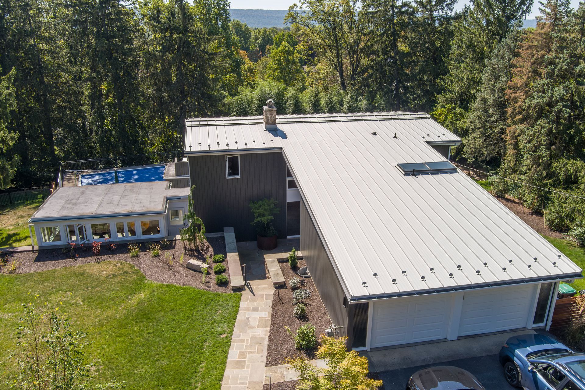 Aerial view of a gray house with a metal roof, a sunroom extension, a swimming pool, and a stone walkway in a wooded area.
