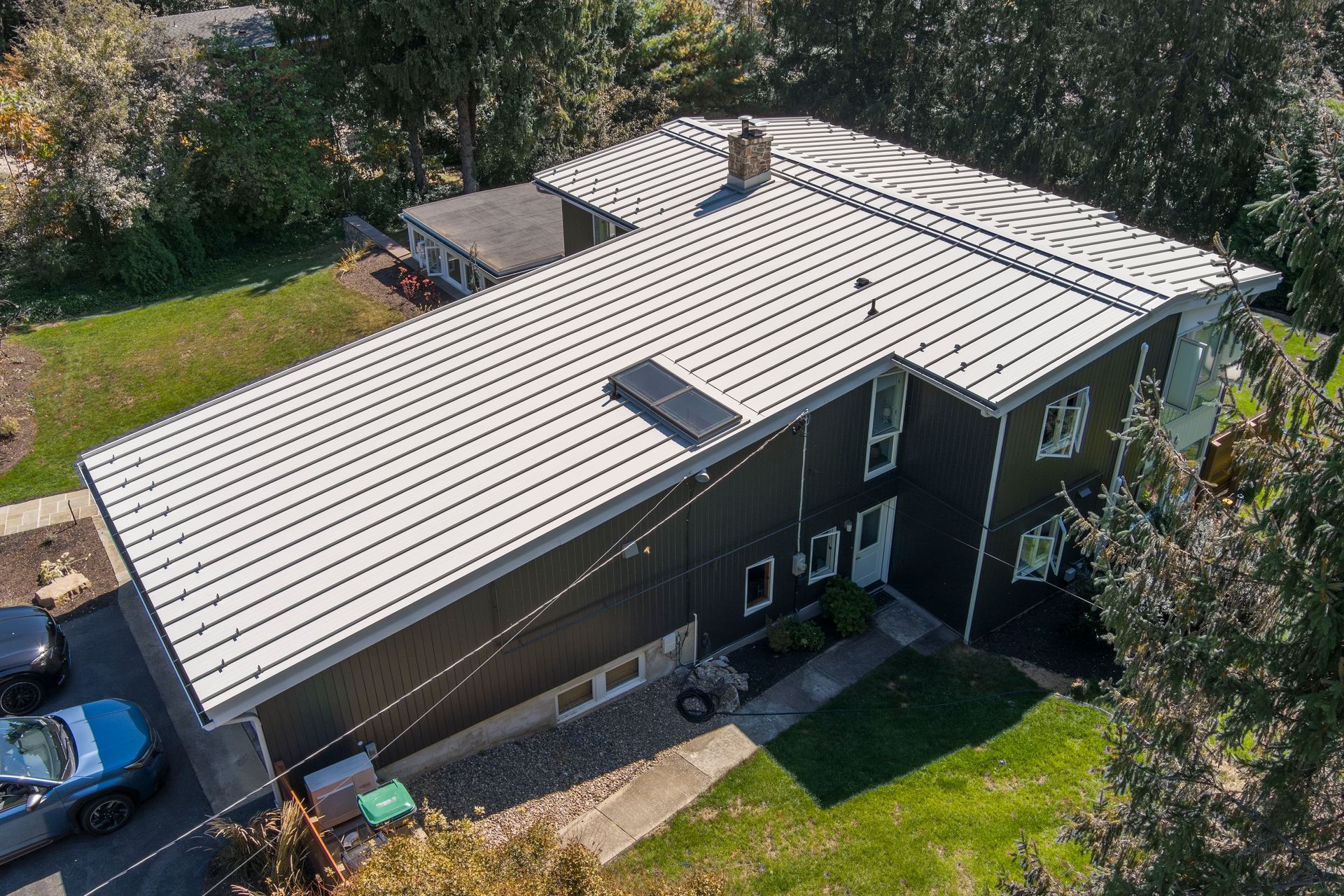 An aerial view of a dark-sided home with a prominent white metal roof, surrounded by green trees and a grassy yard.