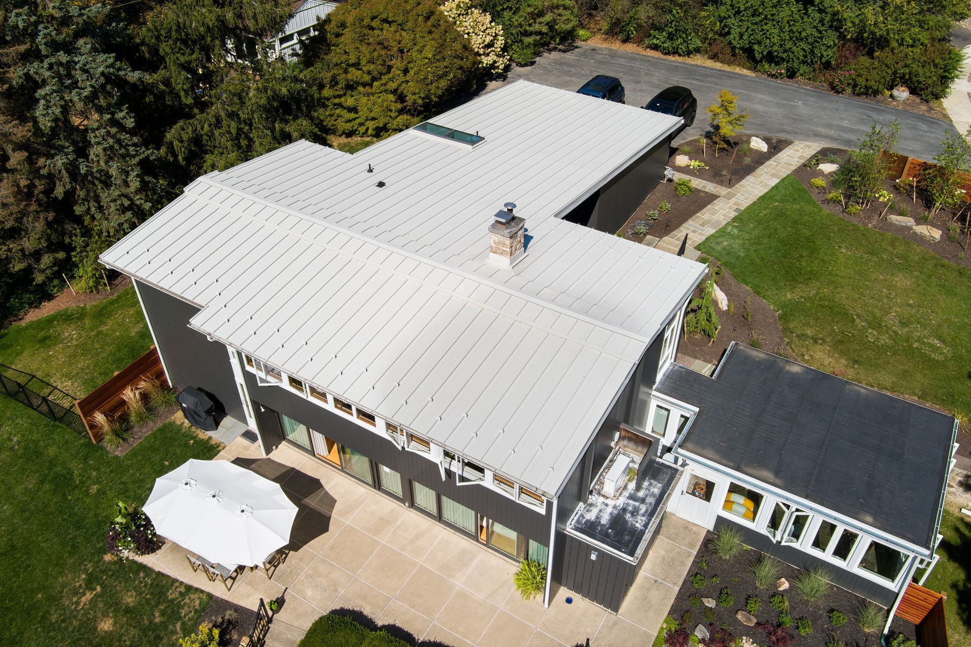 Aerial view of a house with a gray metal roof, a dark side extension, a back patio, and a white umbrella on the lawn.