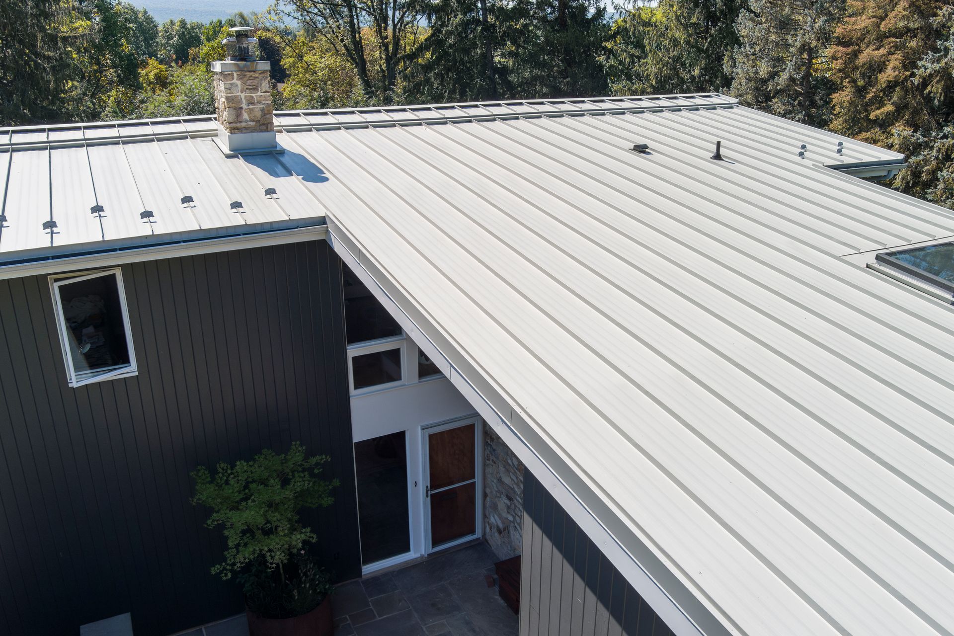 An aerial view of a house with a modern light-gray metal roof, stone chimney, and dark siding surrounded by green trees.