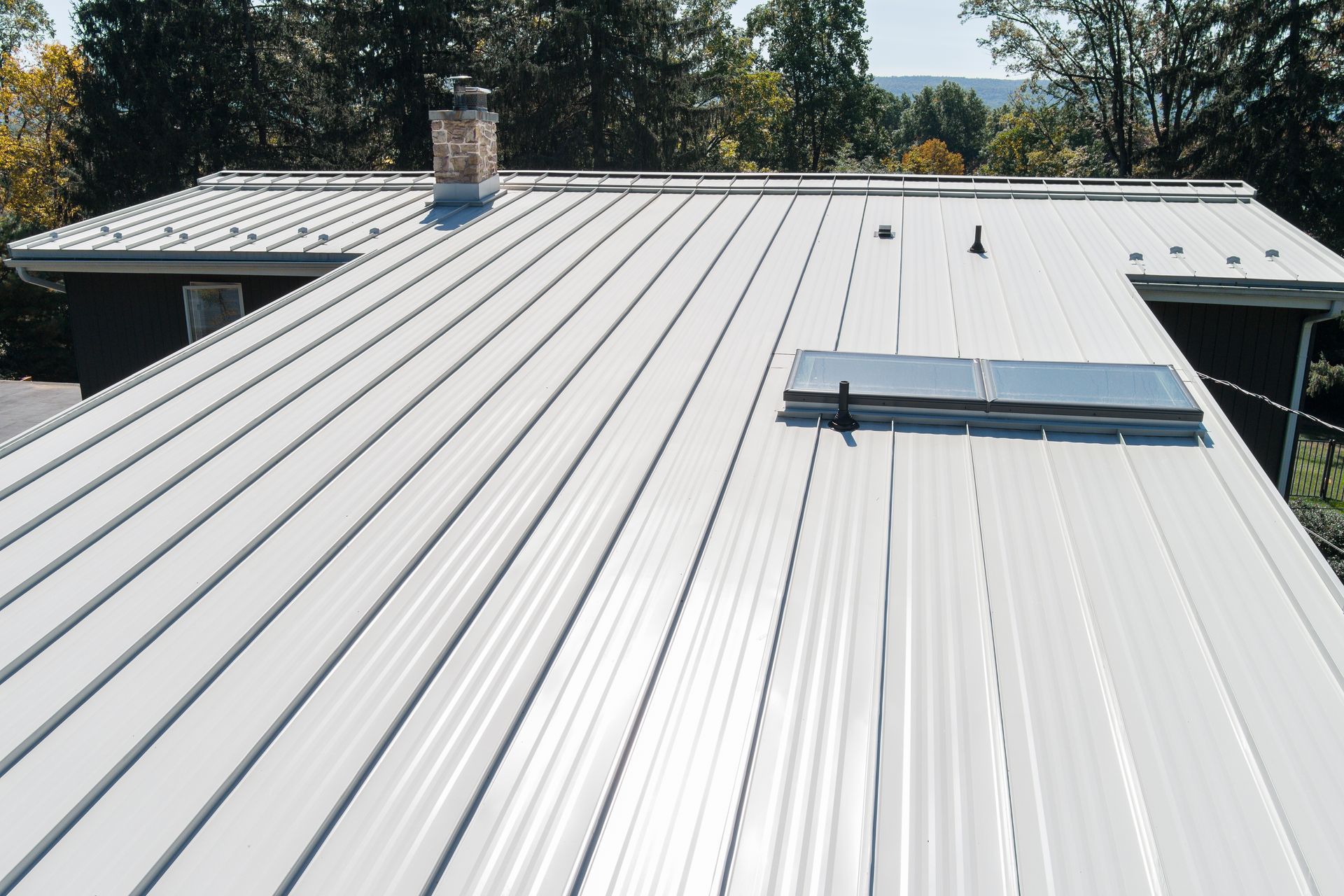 A high-angle view of a white, ribbed metal roof on a residential home with a chimney and a rectangular skylight.