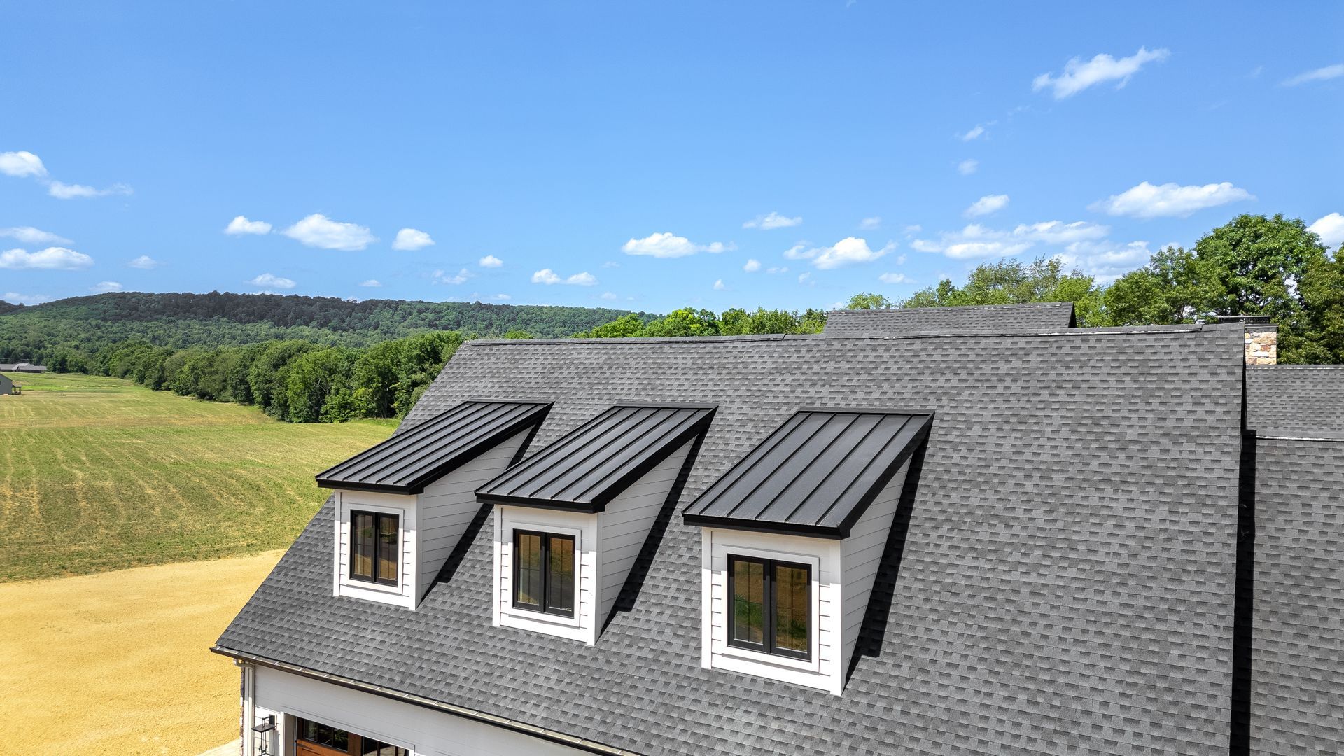 Three dormers with black metal roofs on a gray shingled roof against a blue sky and green trees.
