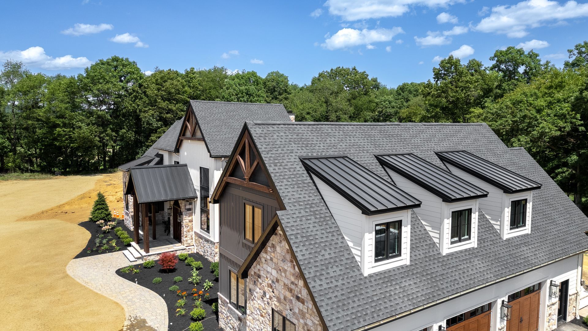 A modern house with a gray roof, stone facade, and three dormer windows against a green tree background.