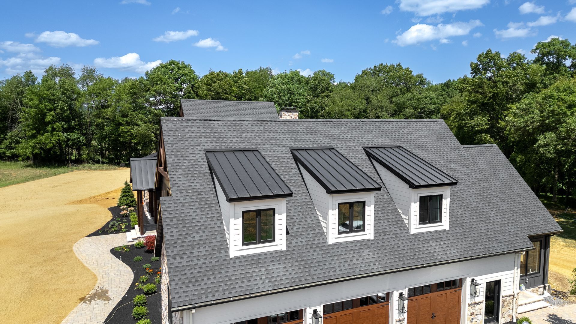 House with gray roof, white siding, and three black dormers. Brown garage doors, green trees in background.