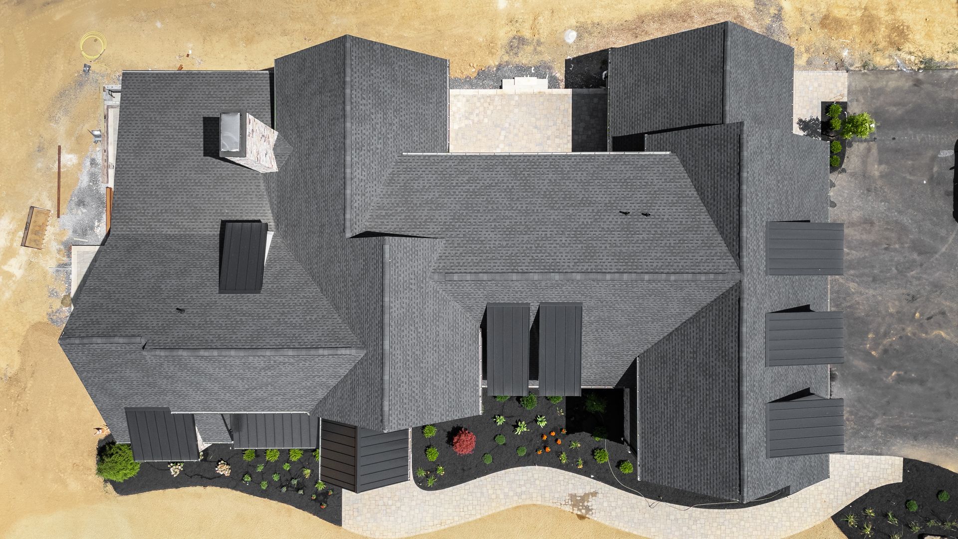 Overhead view of a modern house with a dark gray shingled roof, surrounded by landscaping and a paved driveway.