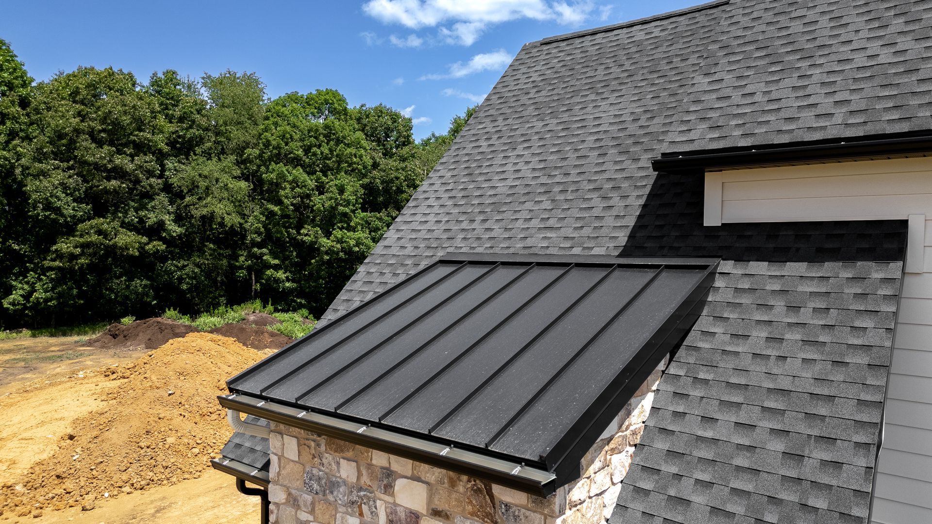 Black metal roof on a dormer, next to a gray shingle roof, with trees and blue sky in the background.