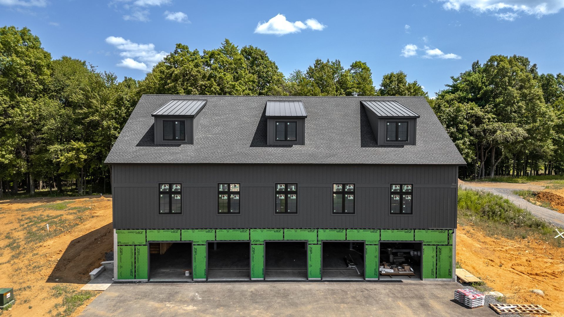 Black building with green garage doors, three dormers, and a treeline backdrop.