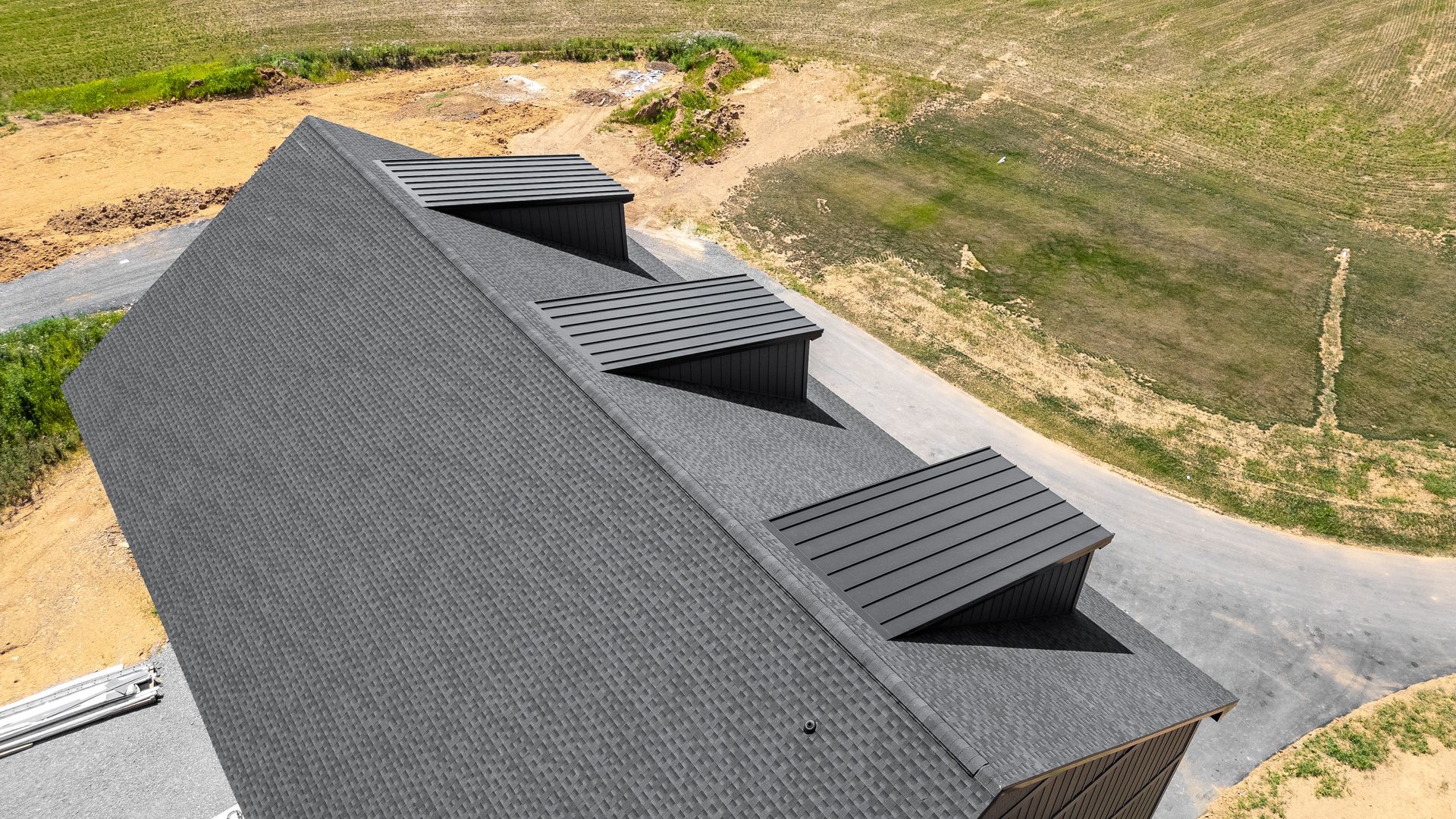 Black shingled roof with three dark vent boxes; gravel driveway, and grassy landscape.