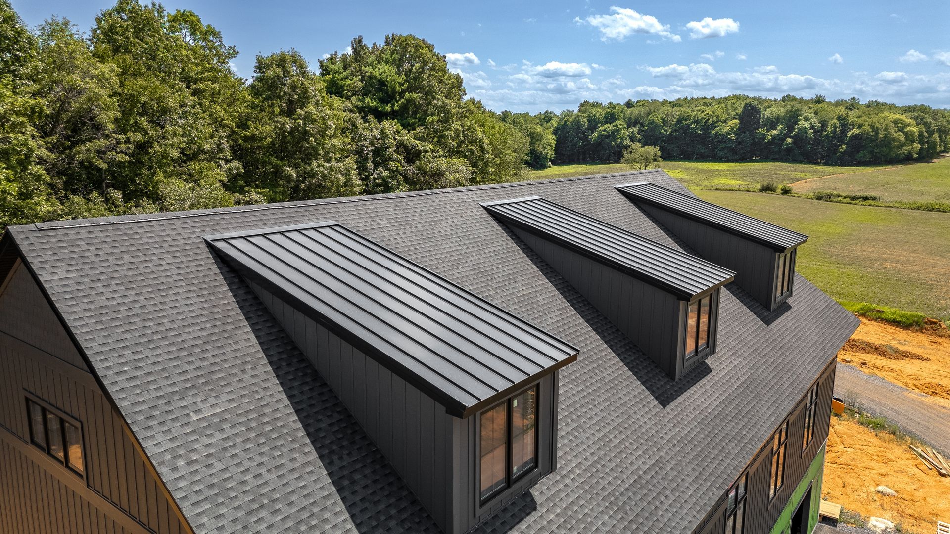 Black roof with three dormers, set against a blue sky and a field.