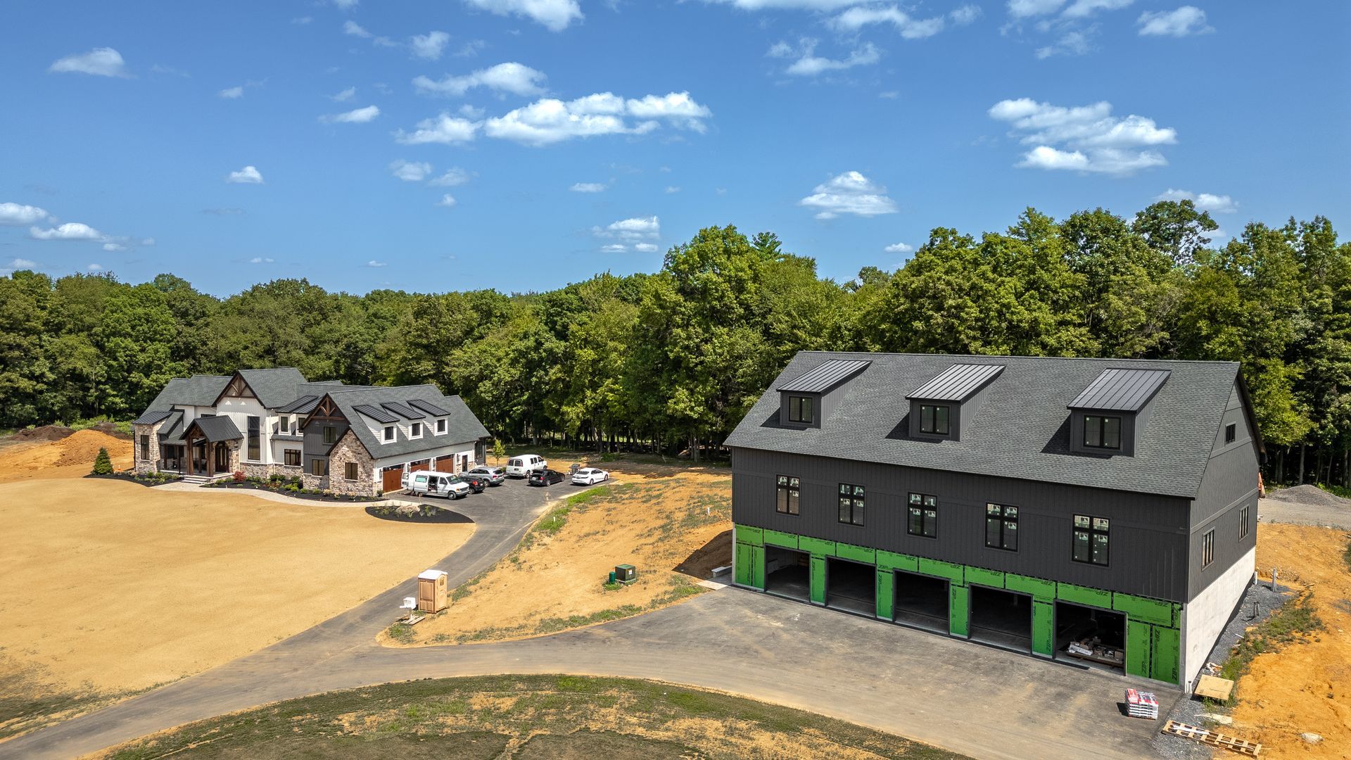 Modern house and garage under construction on a sunny day, surrounded by trees and dirt.