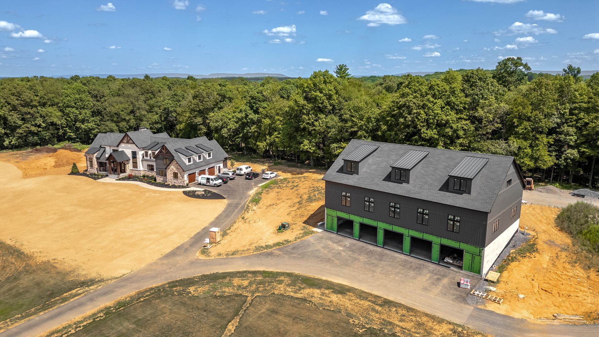 Aerial view of a house and garage on a hilltop with surrounding trees, a driveway, and blue sky.
