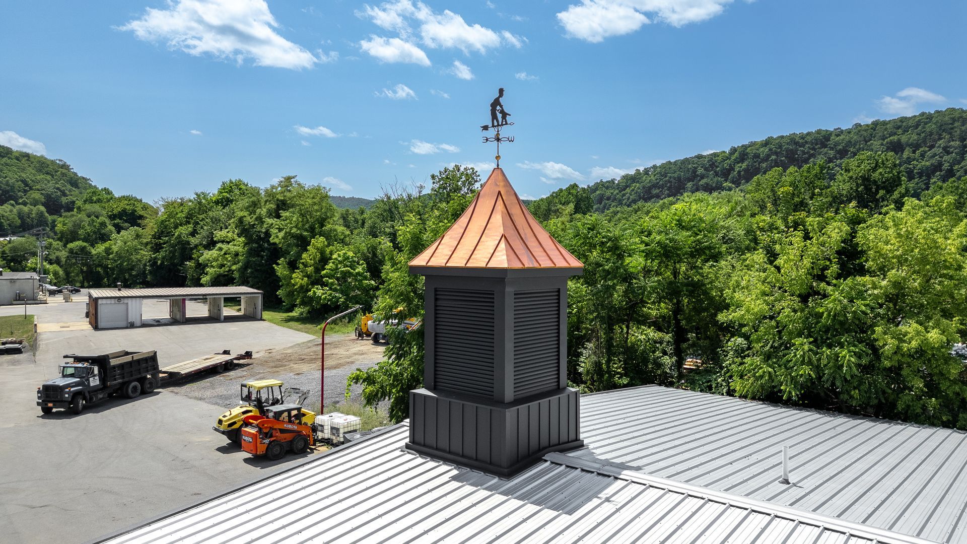 A rooftop structure with a copper roof and a weathervane, set against a backdrop of green trees and a blue sky.
