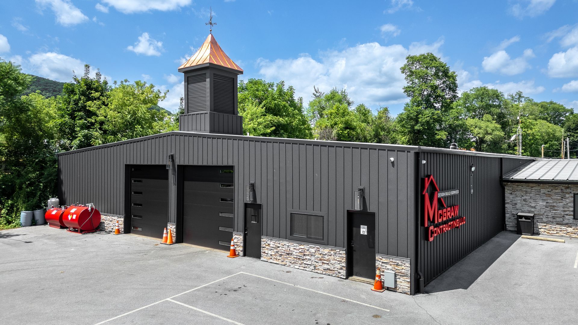 Black metal building with copper-roofed cupola; red logo; two garage doors; stone accents; outdoor setting.