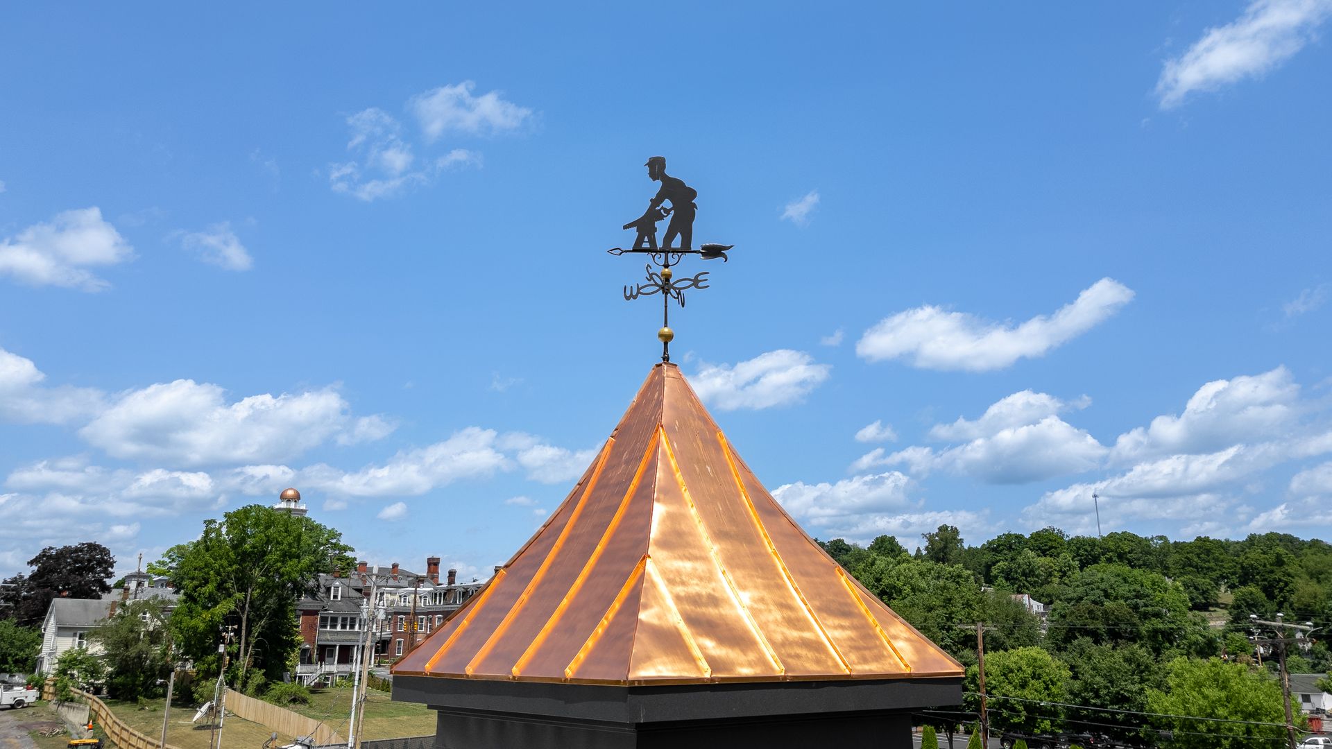 Weather vane atop a copper-clad tower, depicting a silhouetted figure on a roof, blue sky with clouds in background.