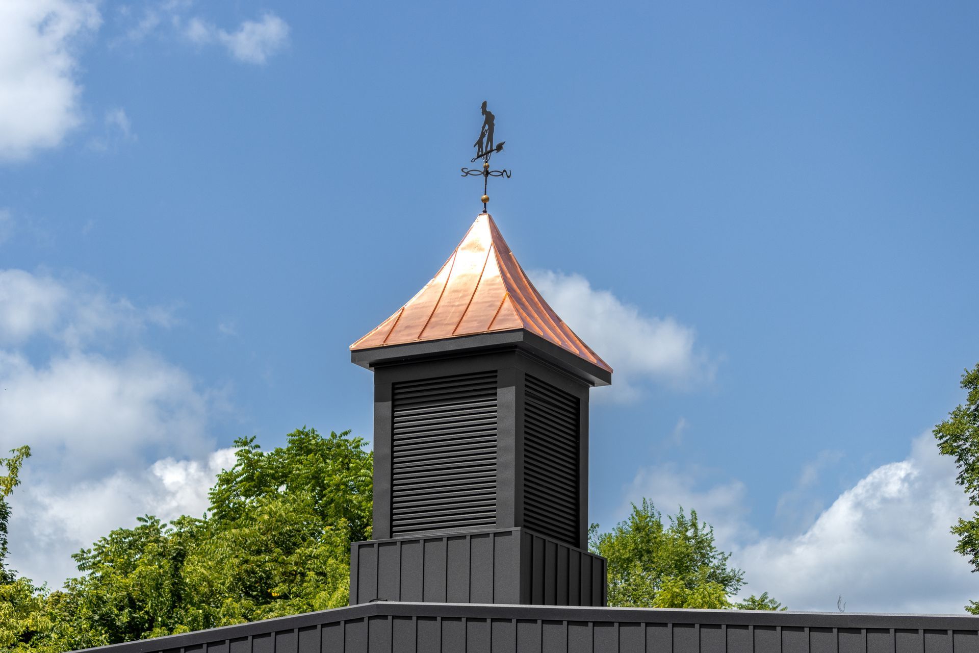 Black cupola with copper roof, weather vane, and slatted sides, against a blue sky.