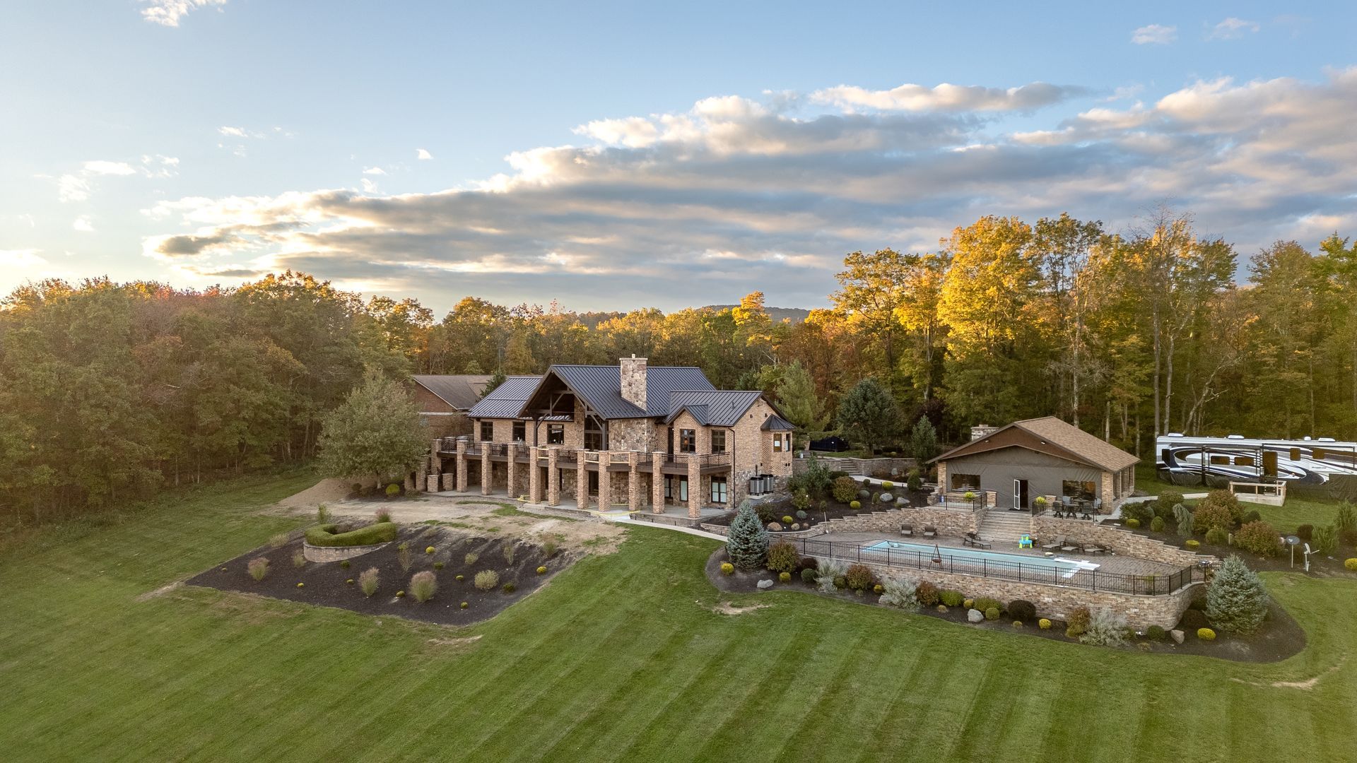 Luxurious stone house with pool on a vast lawn, surrounded by trees under a cloudy sky.