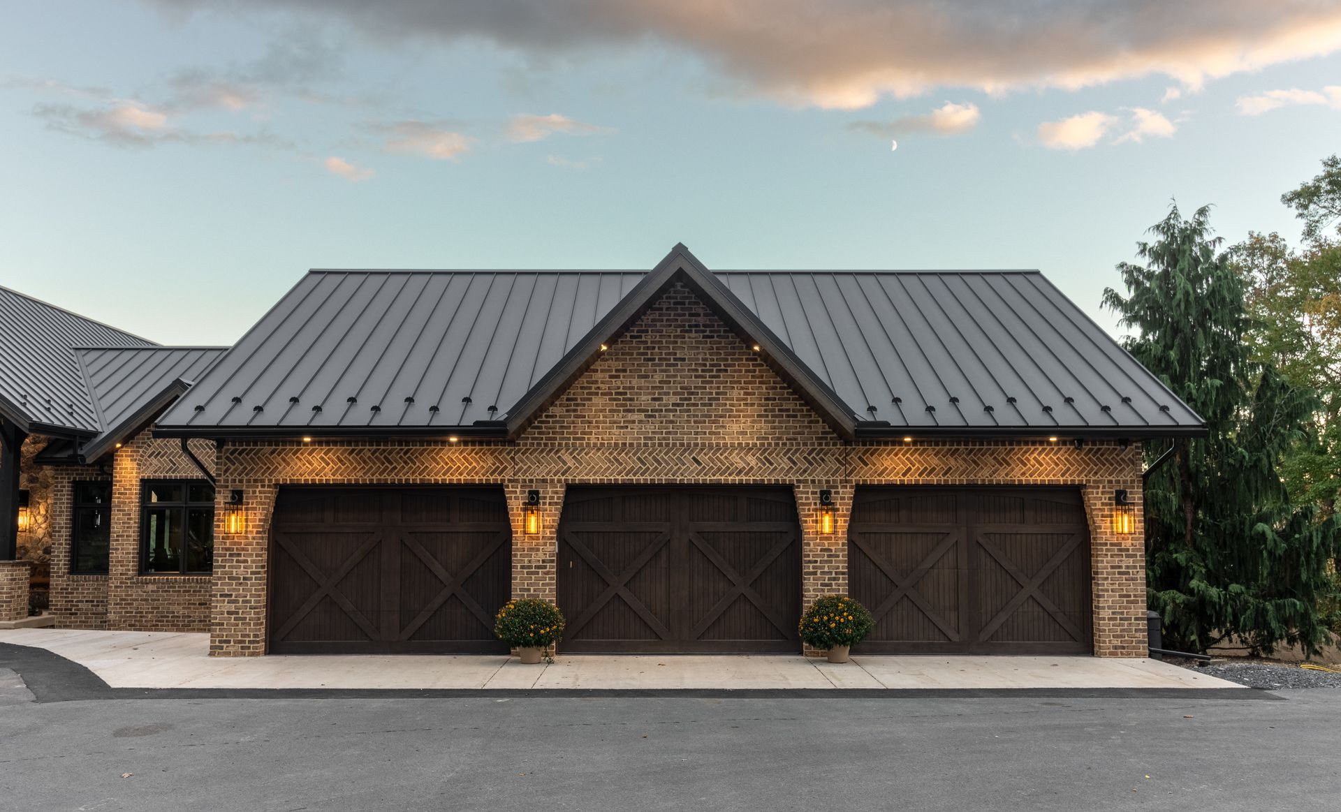 Three-car garage with brown doors, brick facade, dark roof, and accent lighting, set against a twilight sky.