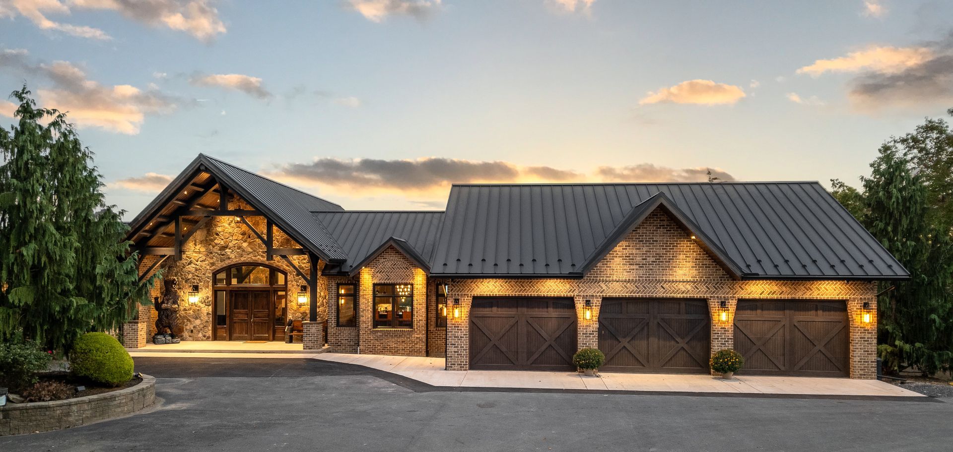 A rustic, stone and wood home with a three-car garage against a sunset sky.