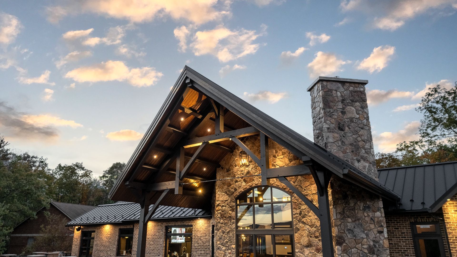 Stone lodge with dark wood accents, stone chimney, and large arched window, under a cloudy sky.