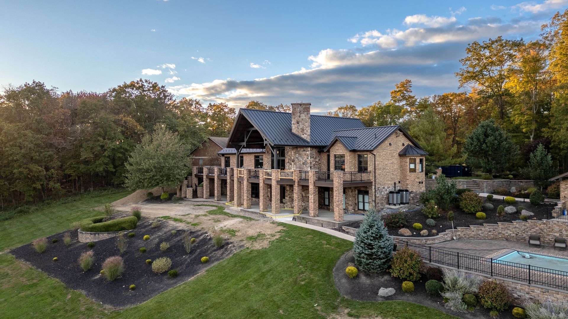 Stone mansion with columns, pool, and landscaping, set in a wooded area under a blue and cloudy sky.