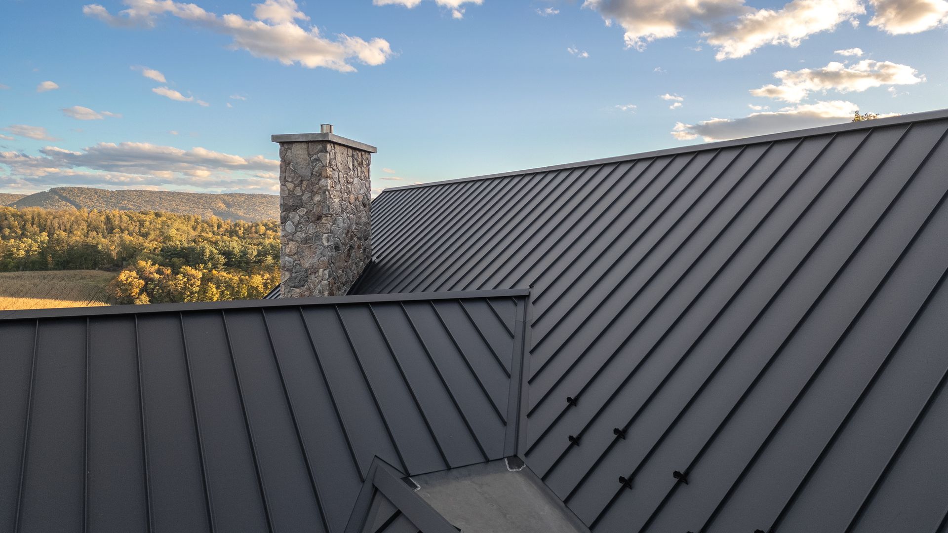 Gray metal roof of a house with a stone chimney, blue sky and landscape in the background.